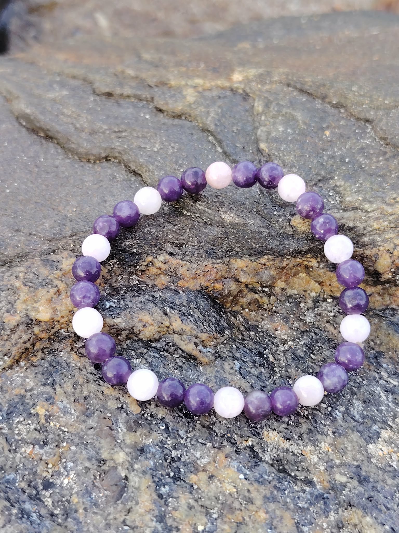 Bracelet Lépidolite Kunzite sur rocher en Bretagne, perles violettes et roses en pierres naturelles, apaisement émotionnel et douceur intérieure.
