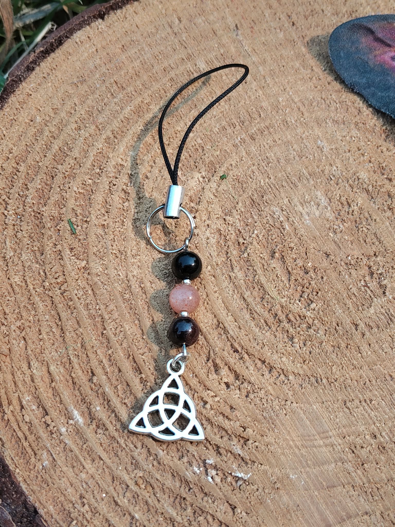 Bijou de téléphone Viking Triquetra accroché sur smartphone, perles noires et rose poudré avec pendentif argenté, harmonie, ancrage et protection.