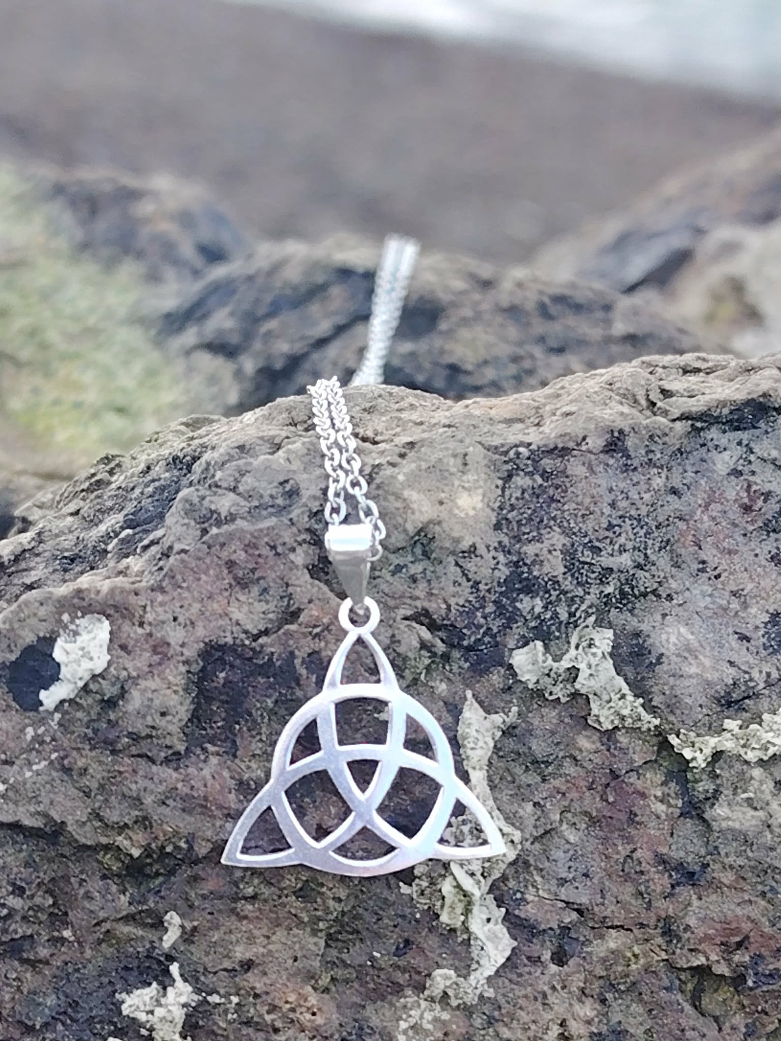 Pendentif Triquetra argent 925 en gros plan sur roche en Bretagne face à la mer, bijou celtique ajouré, ancrage, protection et stabilité intérieure.
