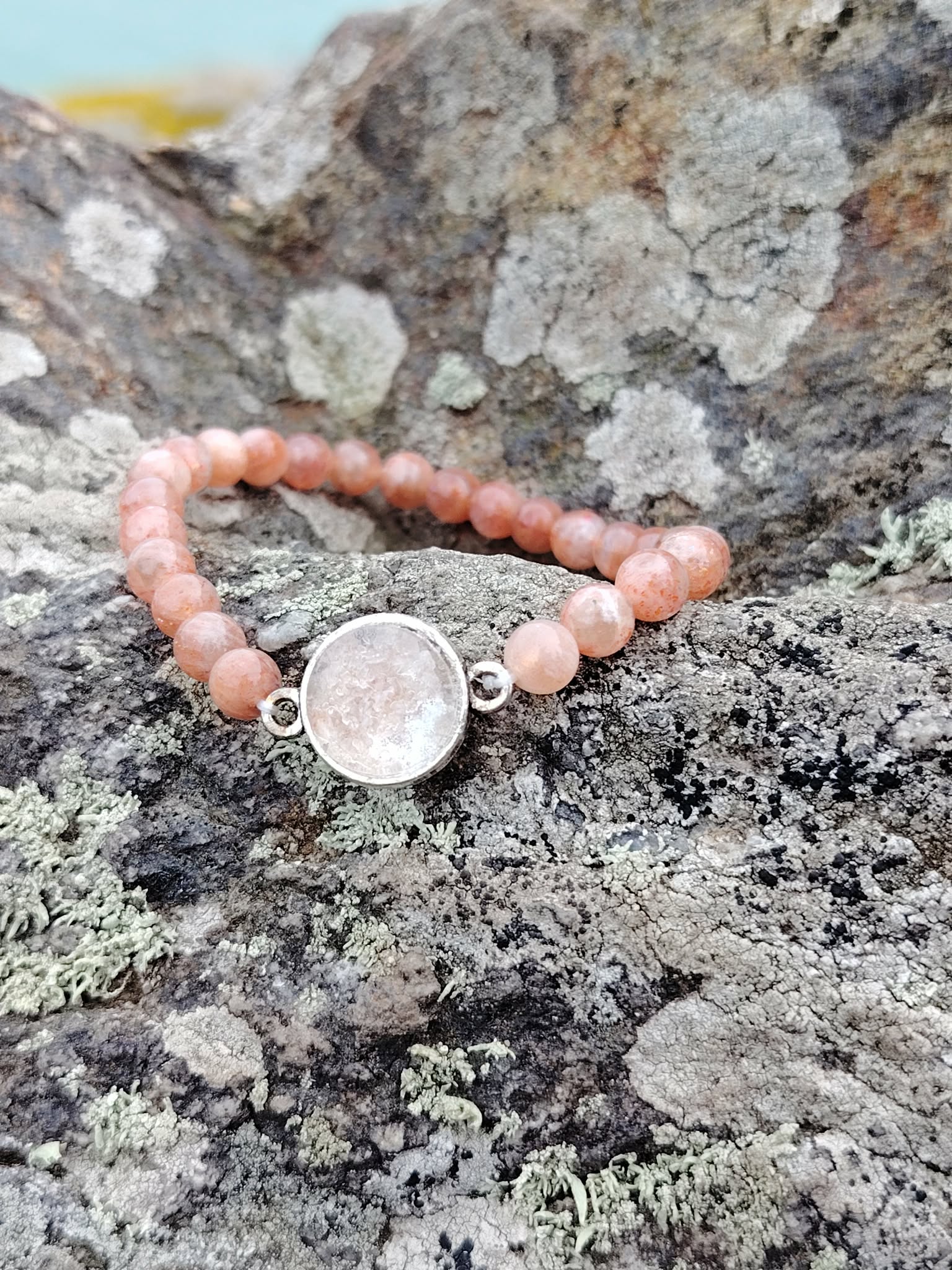 Gros plan bracelet Pierre de Soleil création Magic Gaïa sur rochers en Bretagne, perles rondes pêche rosé et support argenté, optimisme et assurance.