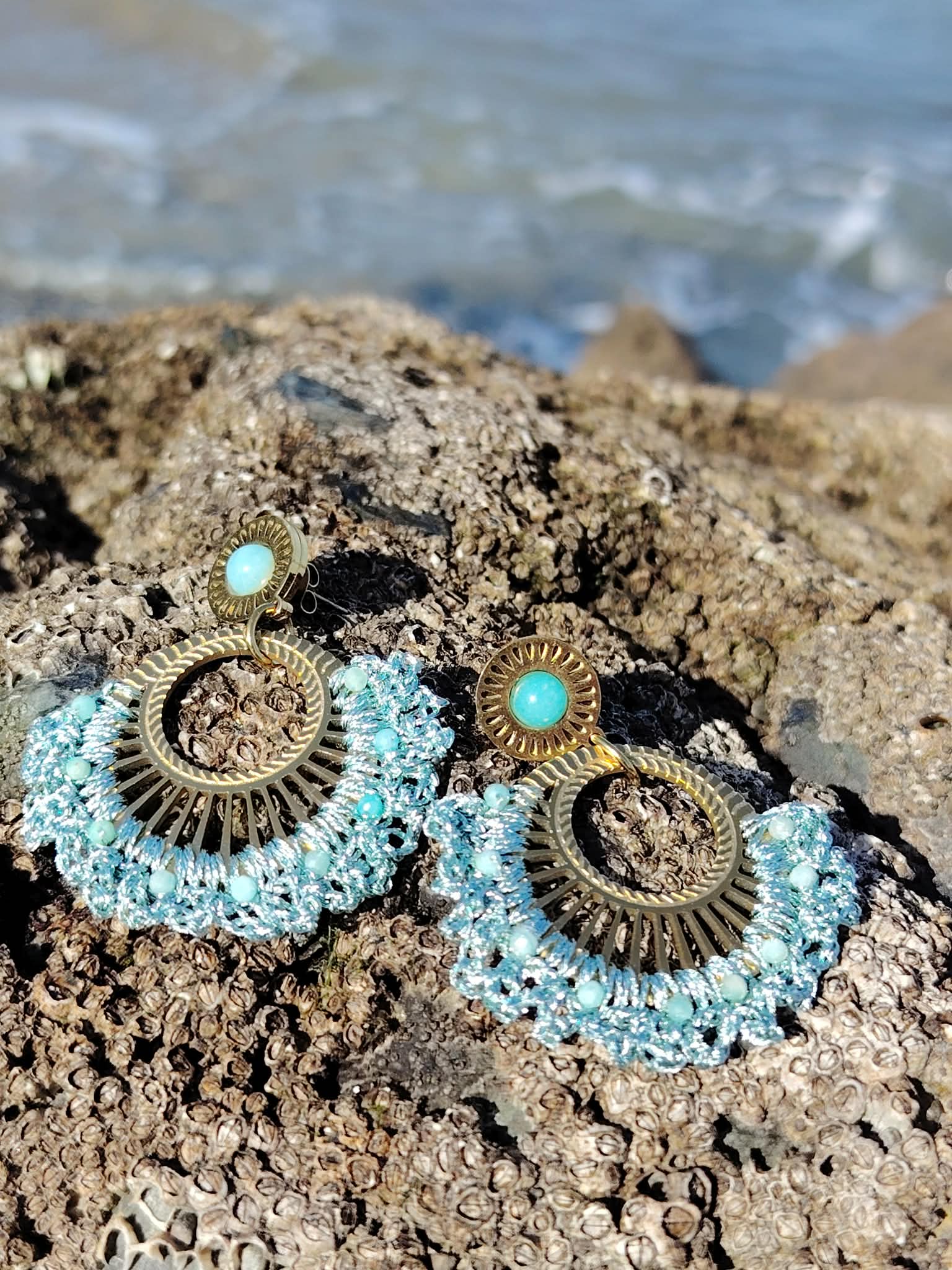 Boucles d’oreilles dorées éventail tissé bleu turquoise posées sur rochers en bord de mer en Bretagne, bijou bohème lumineux