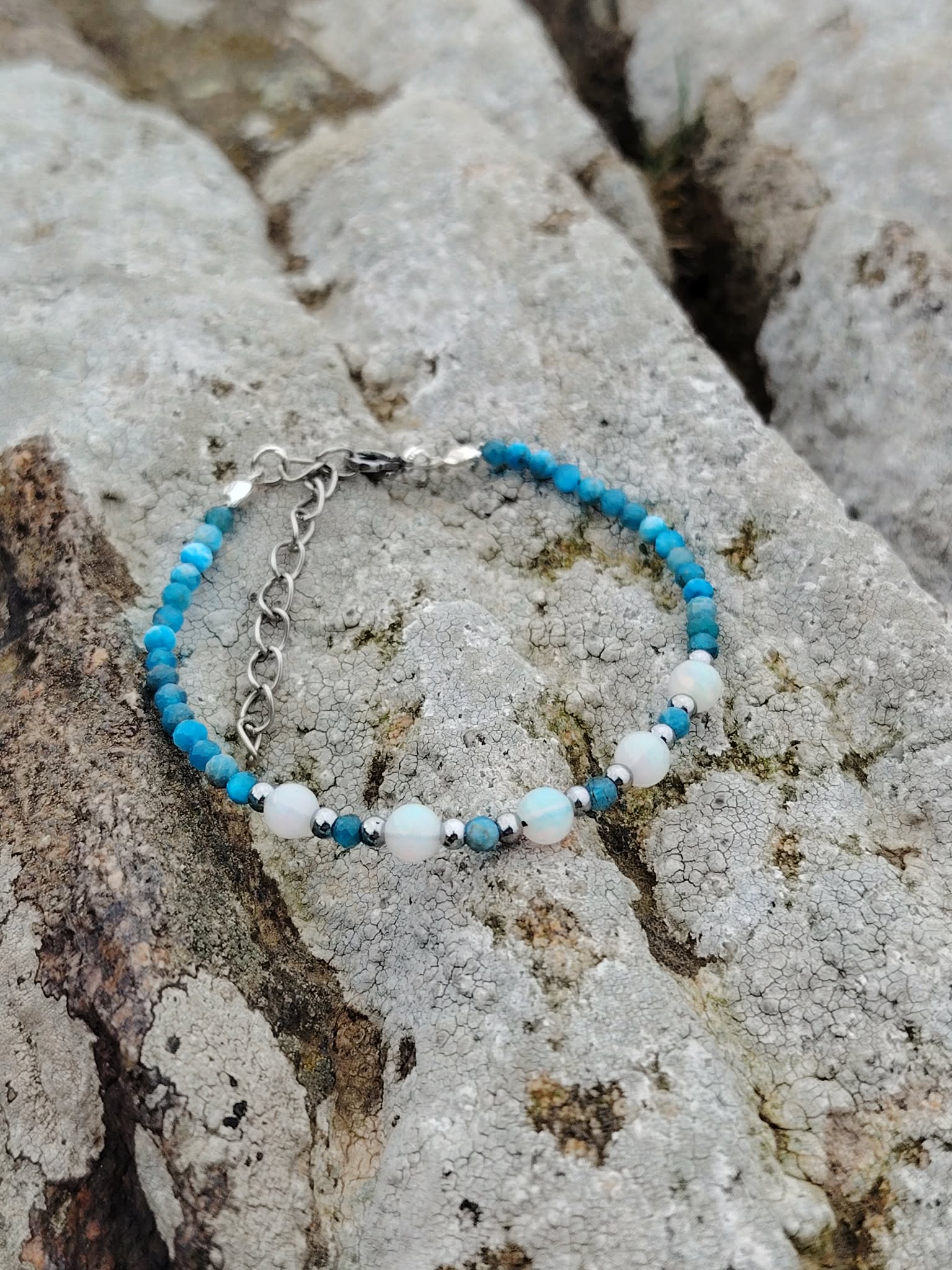 Bracelet fin en apatite facettée et opale sur décor naturel breton, création Magic Gaïa bleue et irisée, parole fluide et intuition.
