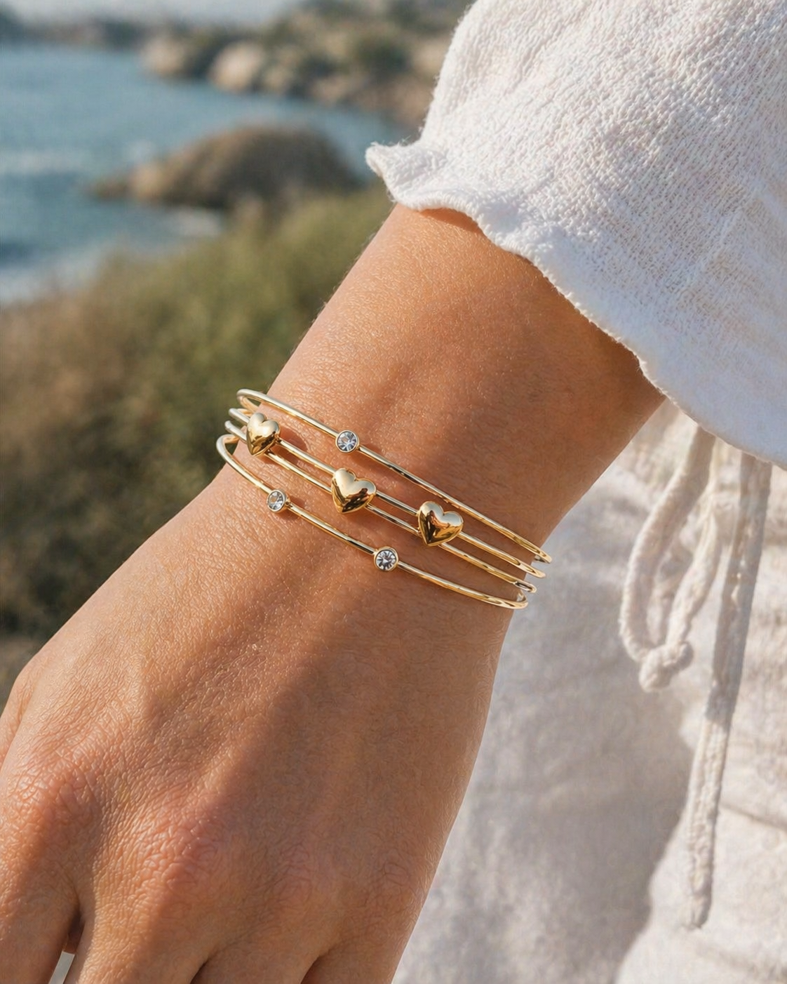 Bracelet jonc doré porté au poignet en lumière naturelle, fils délicats ornés de cœurs et strass, allure chic et solaire.