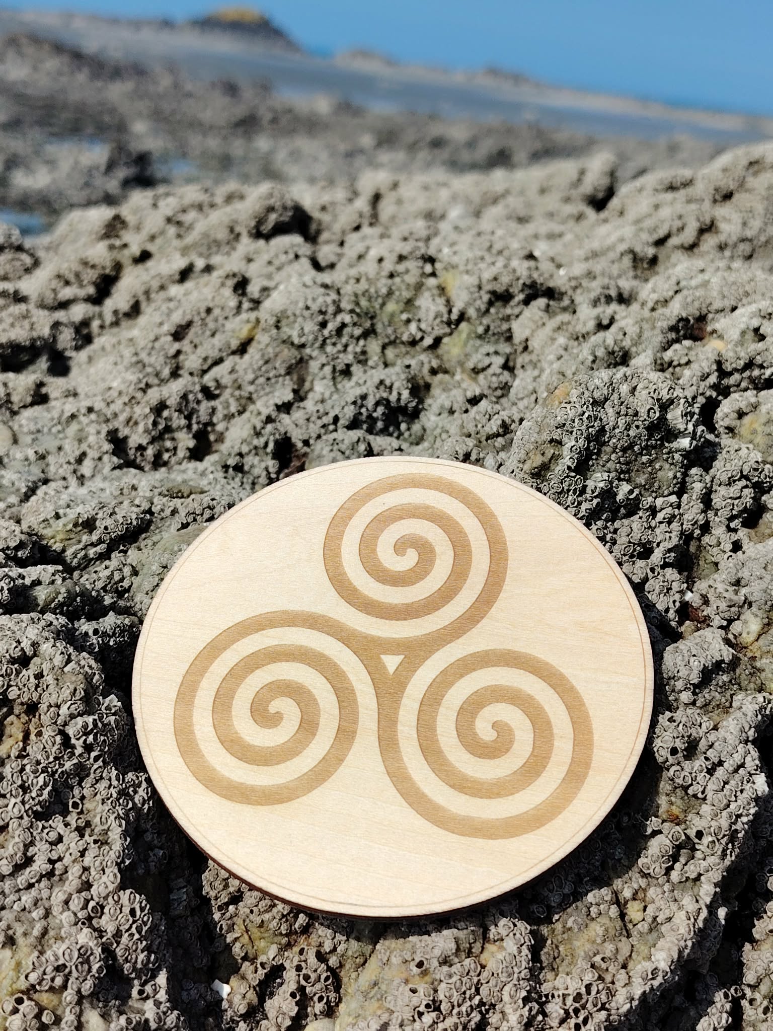 Plaque en bois Triskell 10 cm posée sur rochers en Bretagne, symbole celtique d’équilibre et de cycles naturels.