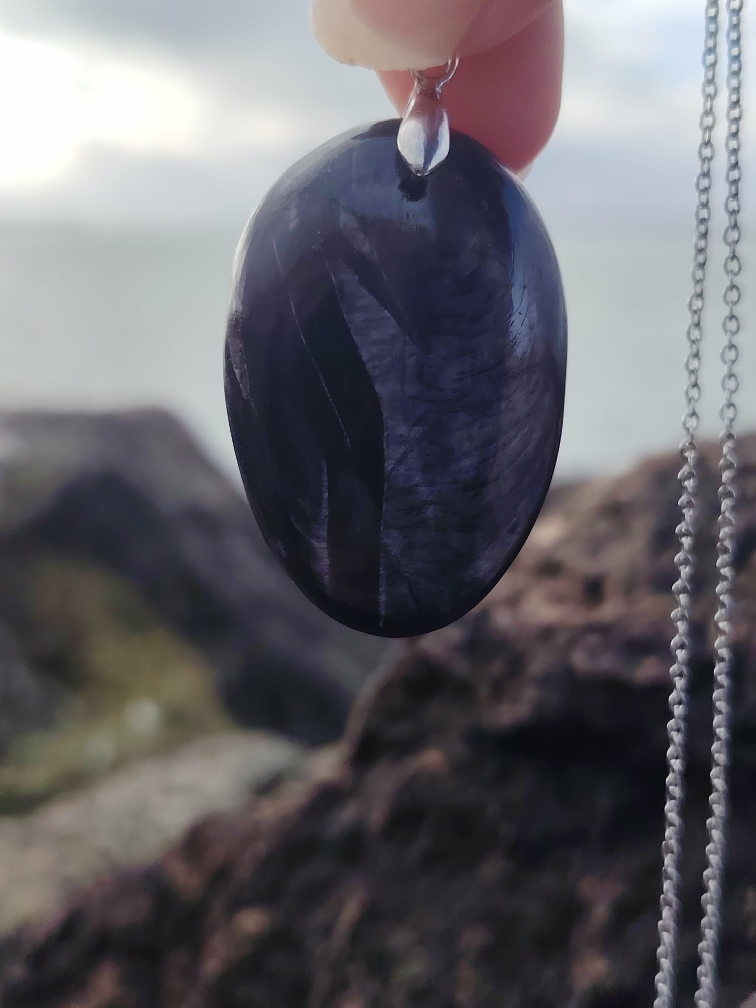 Pendentif Hyperstène tenu à la main devant le littoral breton, pierre noire ovale à reflets argentés, protection symbolique et recentrage