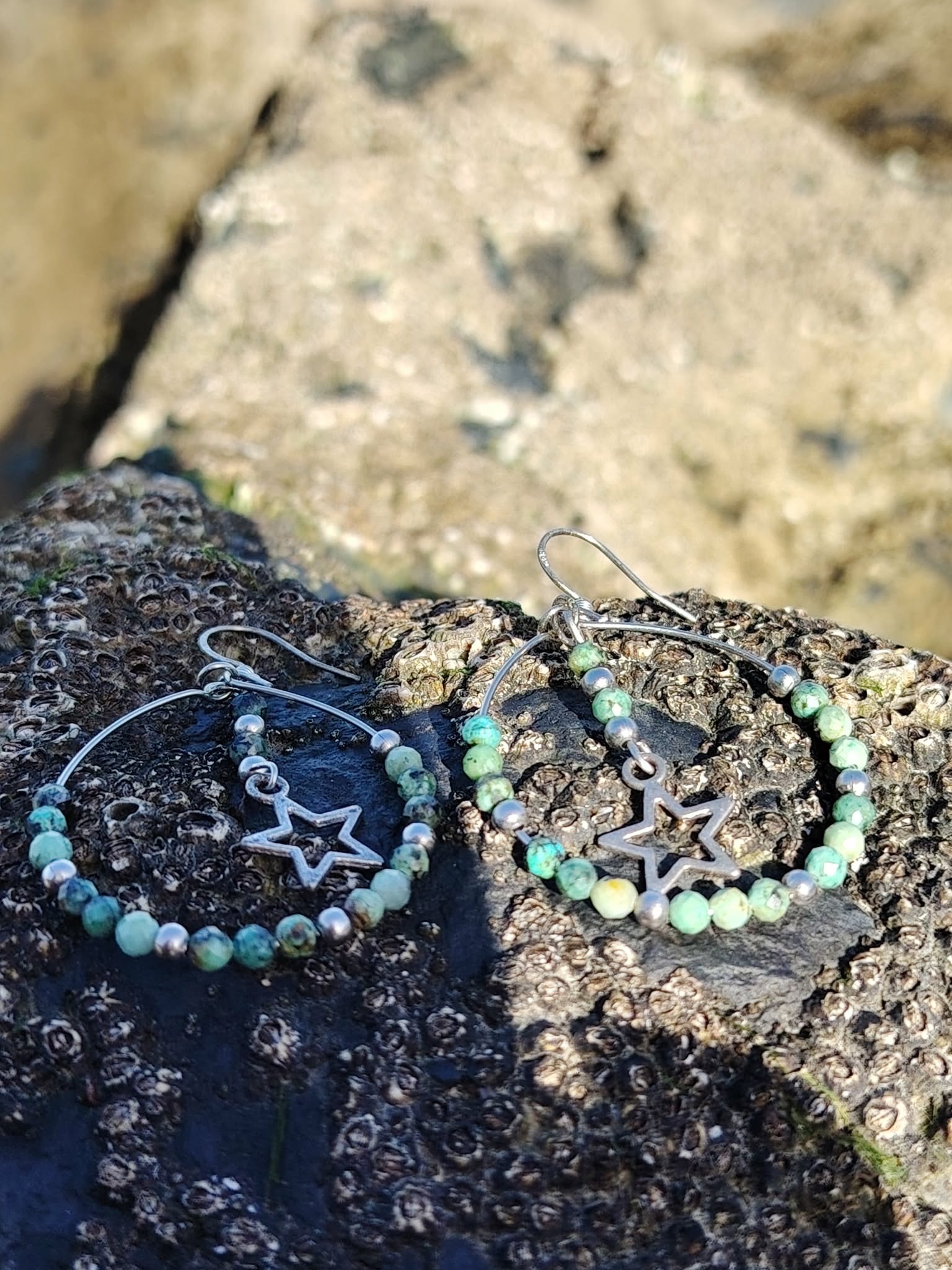 Boucles d’oreilles en turquoise africaine facettée dans un décor naturel breton, créoles en acier inoxydable avec perles marbrées vert bleuté, intercalaires argentés et breloques étoiles.
