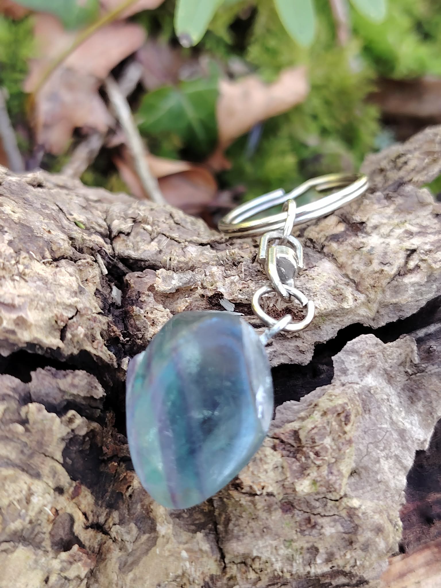 Porte-clés bijou de sac Fluorite multicolore posé sur écorce en forêt, attache argentée et pierre polie verte, bleue et violette.