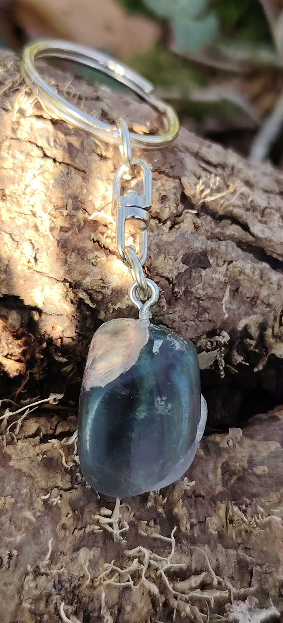 Porte-clés ou bijou de sac Fluorite sur décor naturel, attache argentée et pierre polie aux reflets verts, bleus et violets.