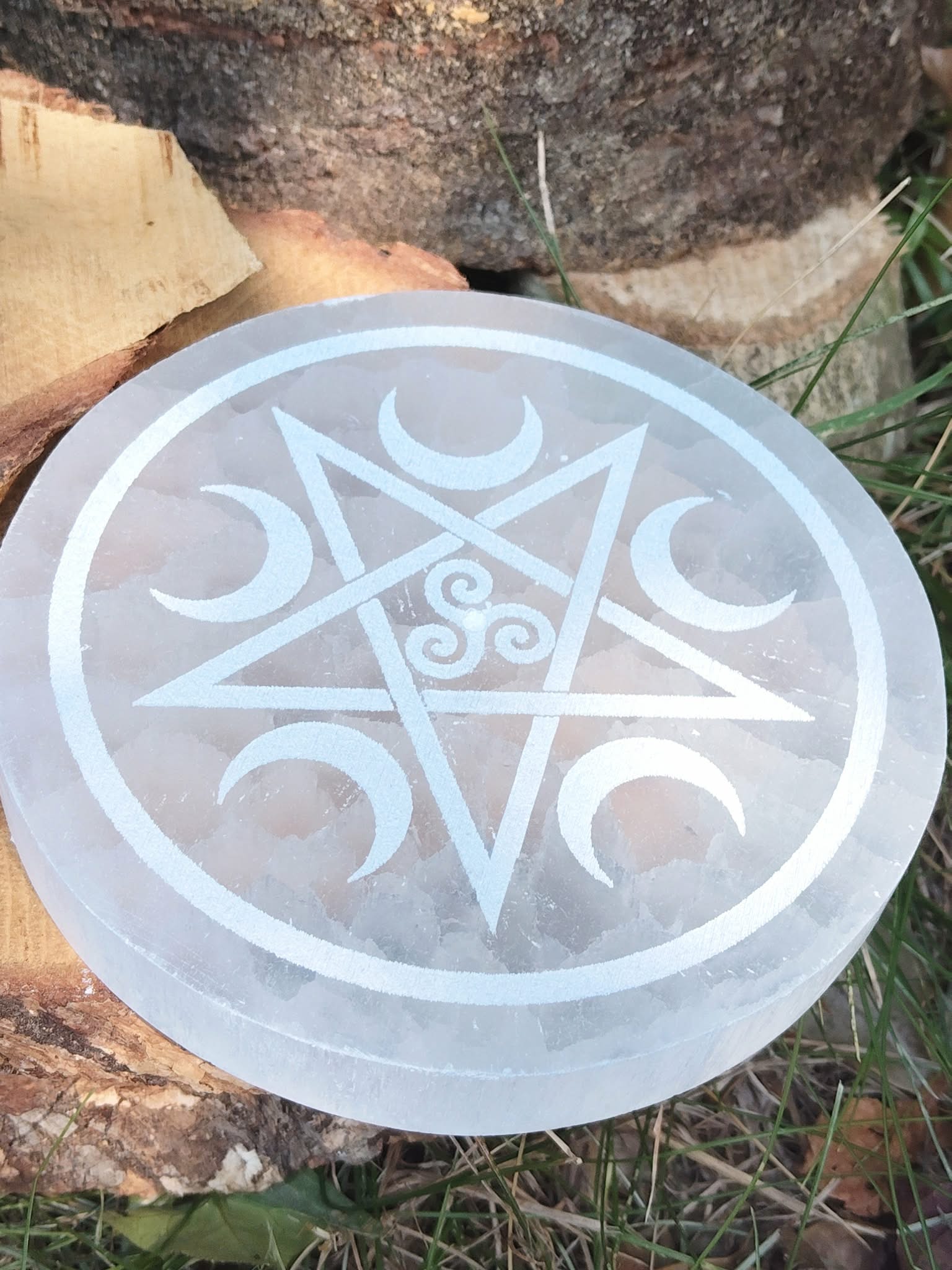 Porte-encens Sélénite Pentacle Lunes et Triskell posé sur rondins de bois et herbe, gravure blanche avec triskell central.