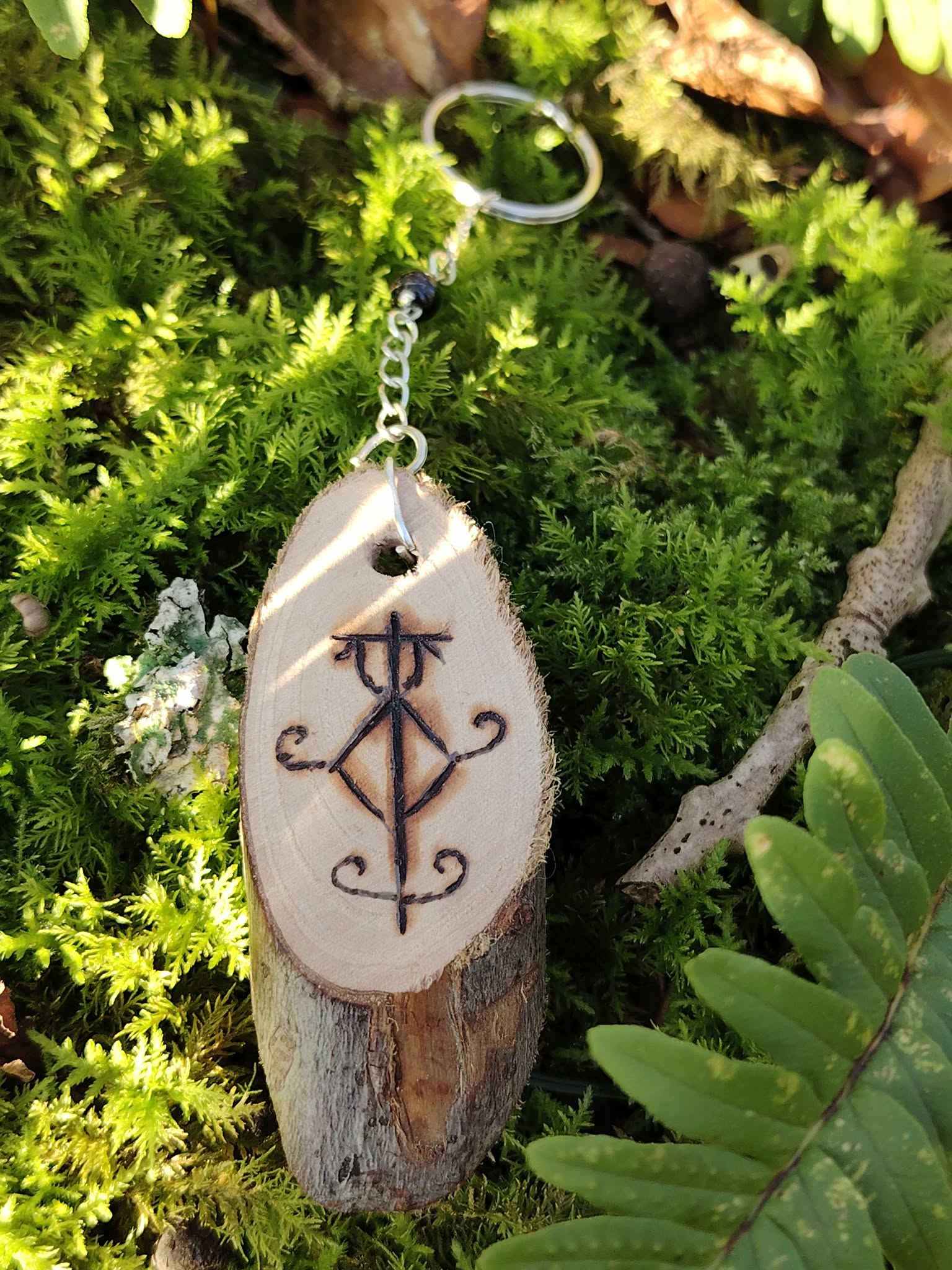 Porte-clés Sigile de Protection Physique posé sur mousse verte avec fougère, bois pyrogravé, chaîne argentée et Tourmaline noire.