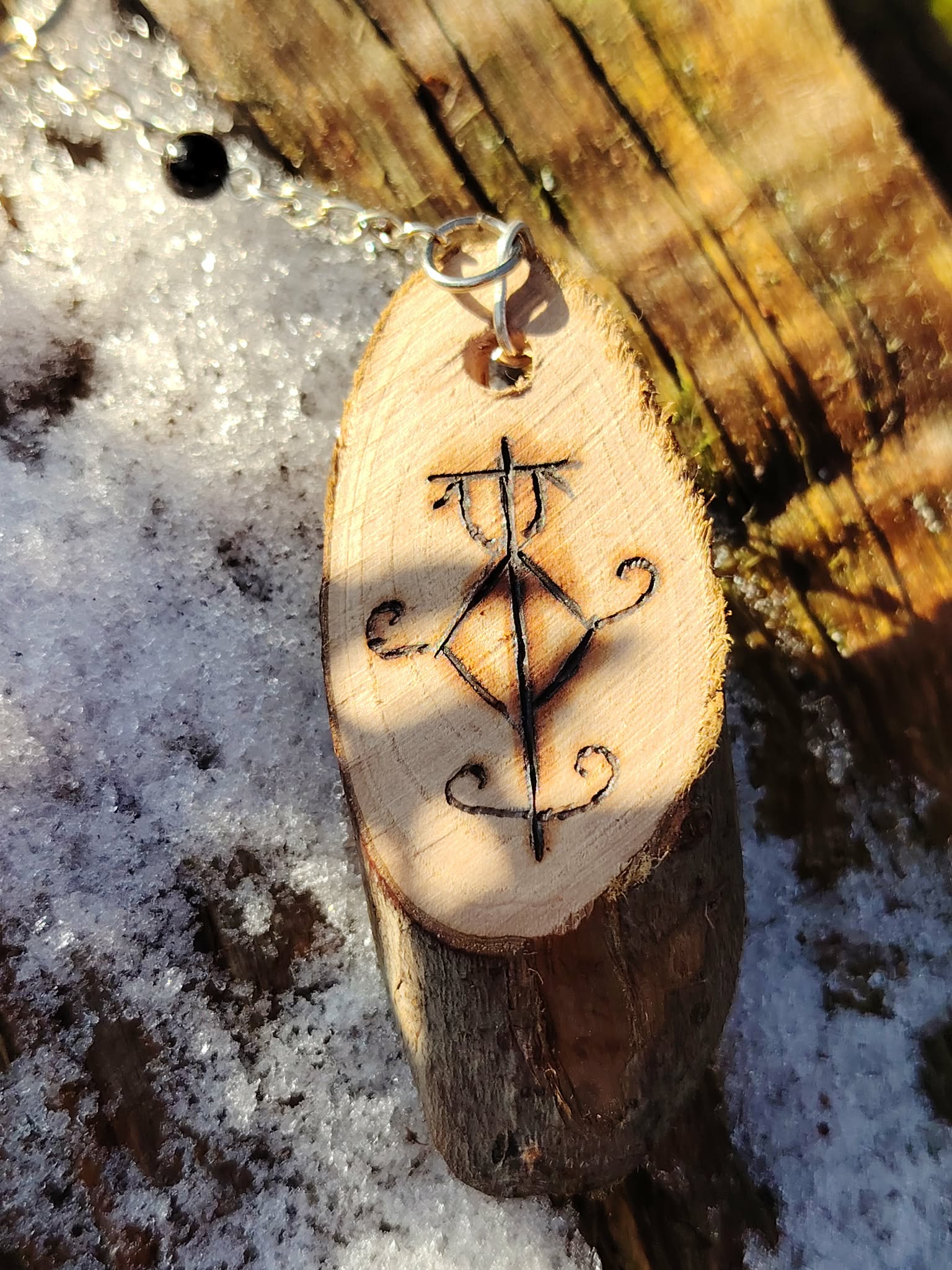 Gros plan sur porte-clés sigile en bois posé sur neige et écorce, symbole pyrogravé, chaîne argentée et Tourmaline noire.
