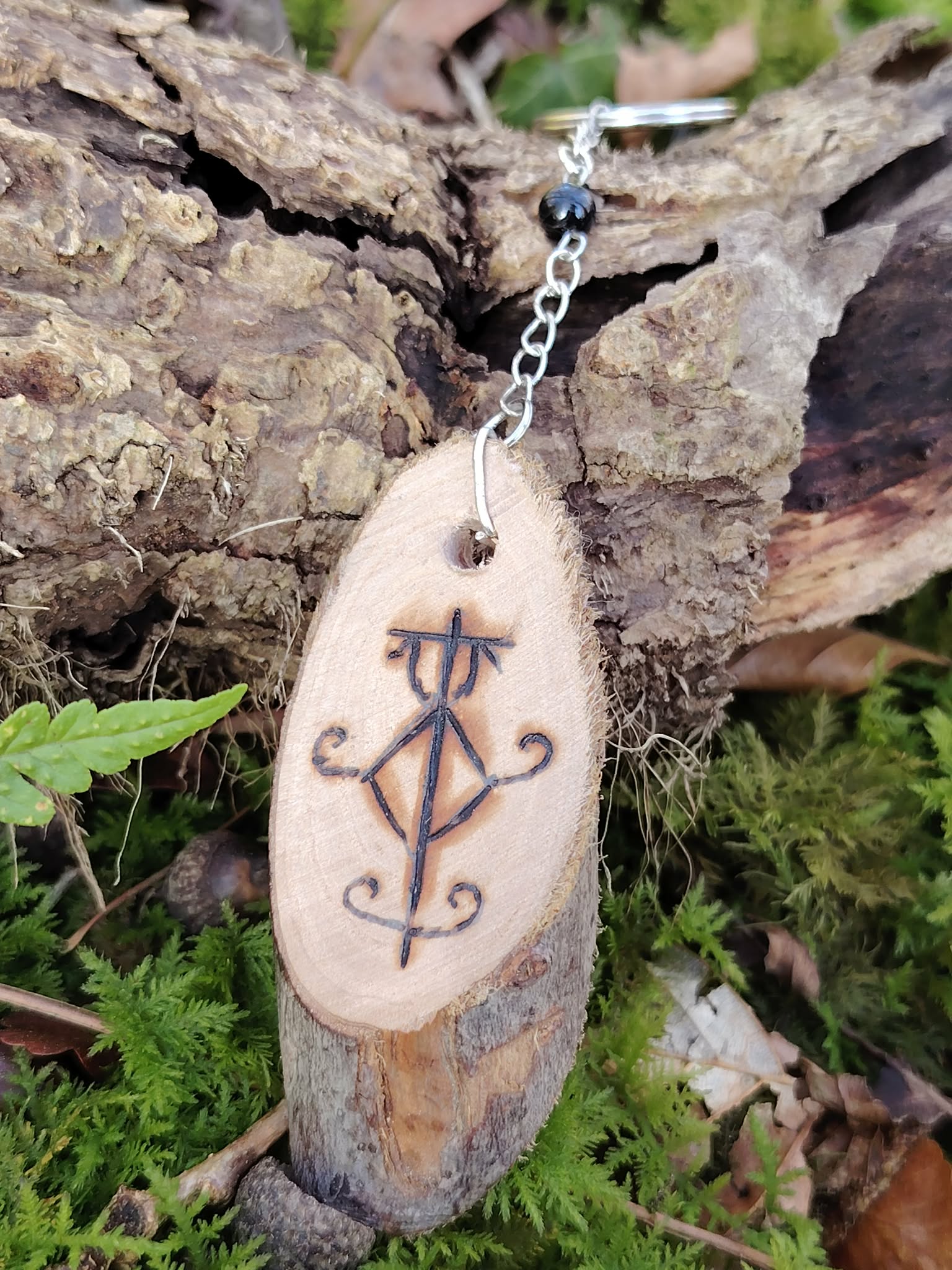 Porte-clés Sigile de Protection Physique en bois pyrogravé, posé contre une écorce avec mousse verte, fougère et Tourmaline noire.