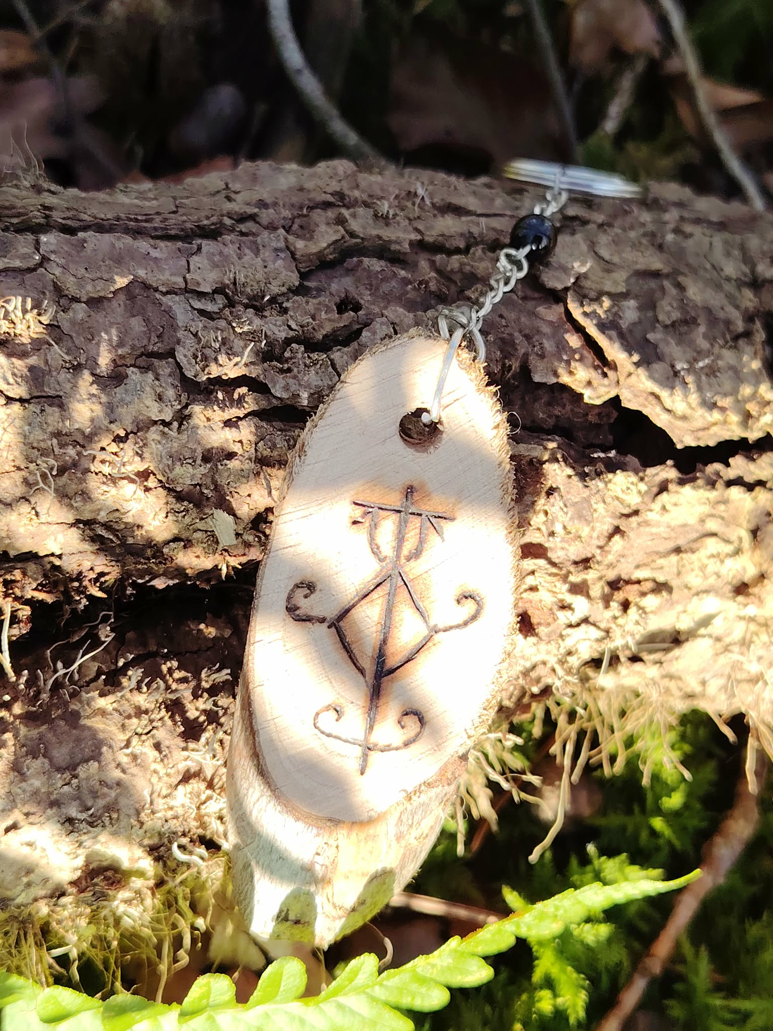 Porte-clés Sigile de Protection Physique suspendu à une écorce en forêt, mousse, fougère et lumière naturelle.