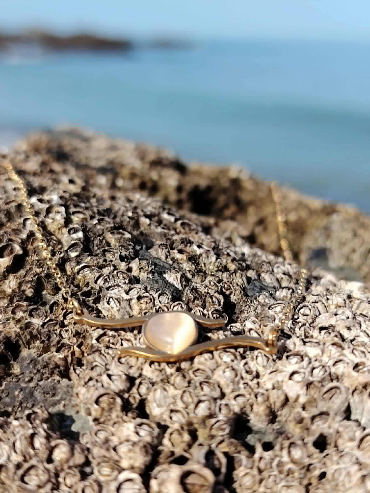 Collier cœur doré rose sur rocher côtier avec mer floutée, pendentif délicat pour amour, douceur et cadeau symbolique.