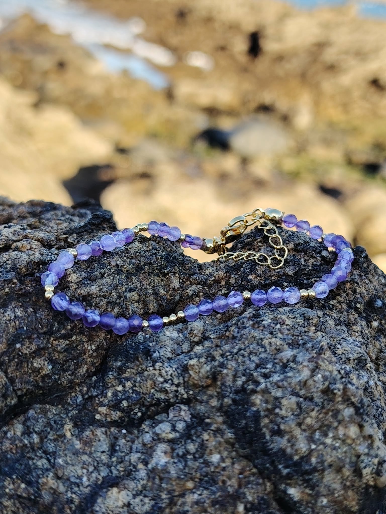 Bracelet améthyste facettée 3 mm posé sur roche en bord de mer, cuivre plaqué or pour apaisement et élégance violette.