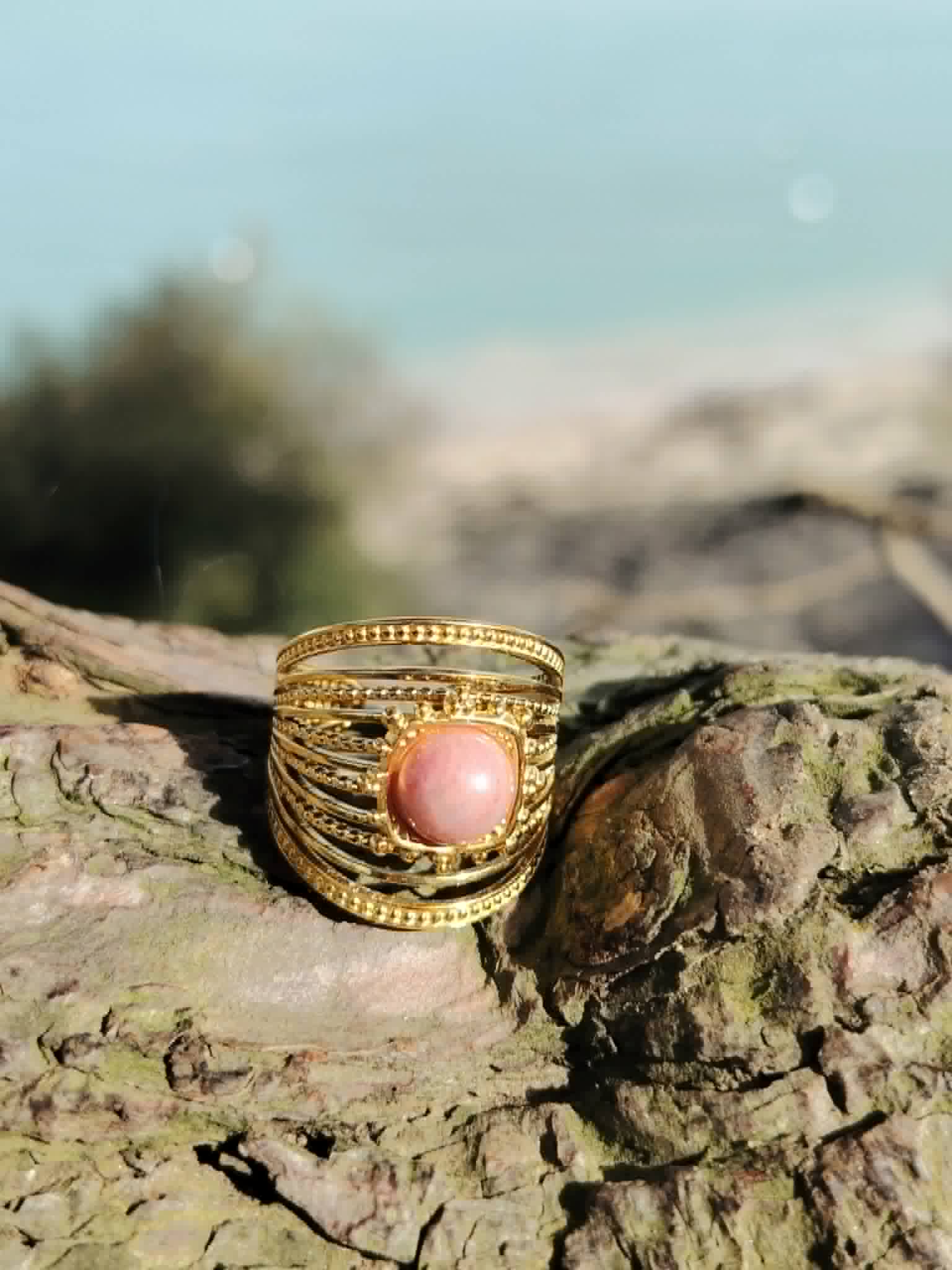 Bague dorée ajustable en acier inoxydable photographiée en Bretagne sur bois flotté, cabochon rose pour douceur et harmonie émotionnelle.