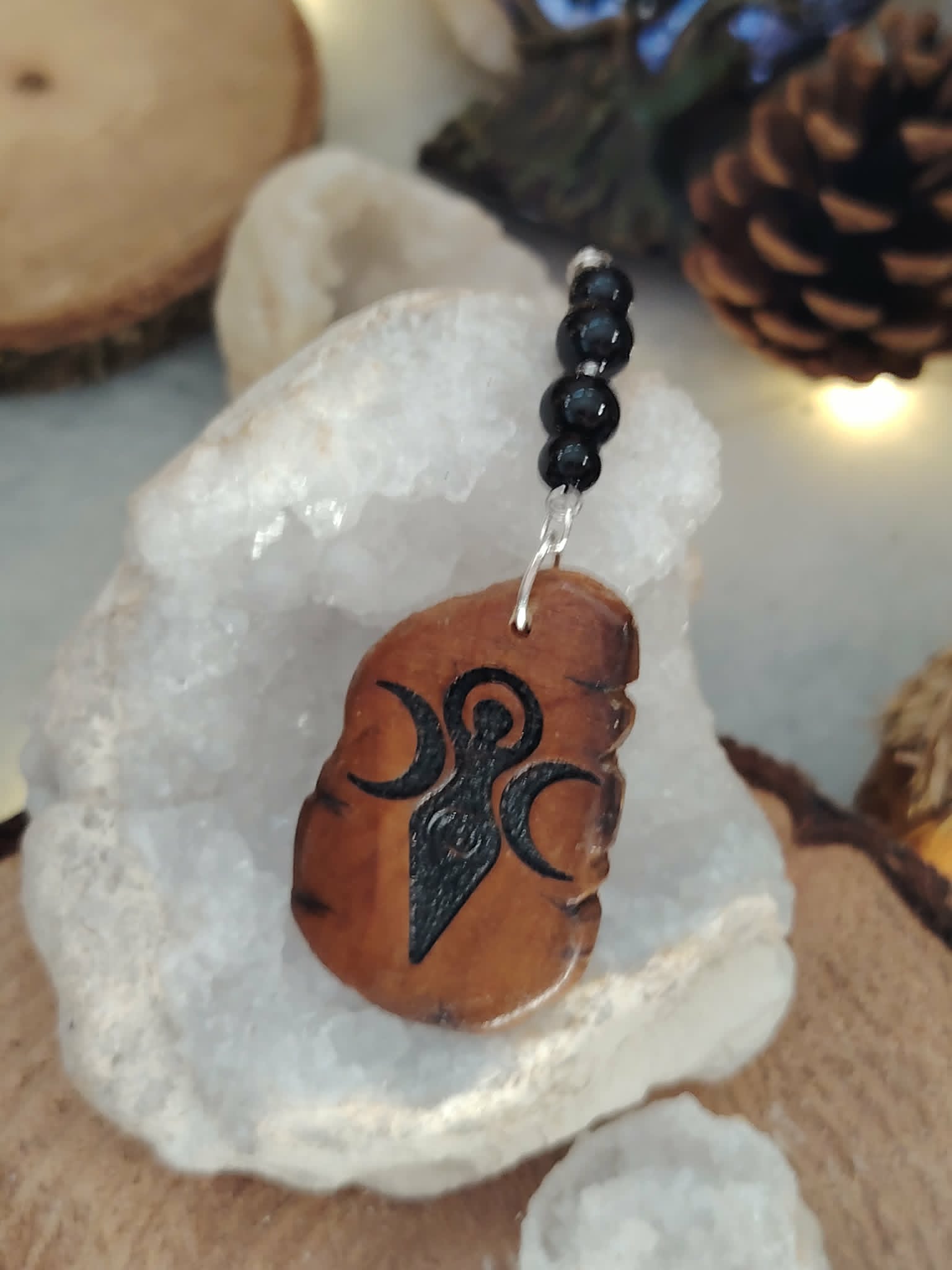 Pendentif de rétroviseur face Gaïa posé sur cristal de roche brut, bois pyrogravé et tourmaline noire dans un décor naturel lumineux.