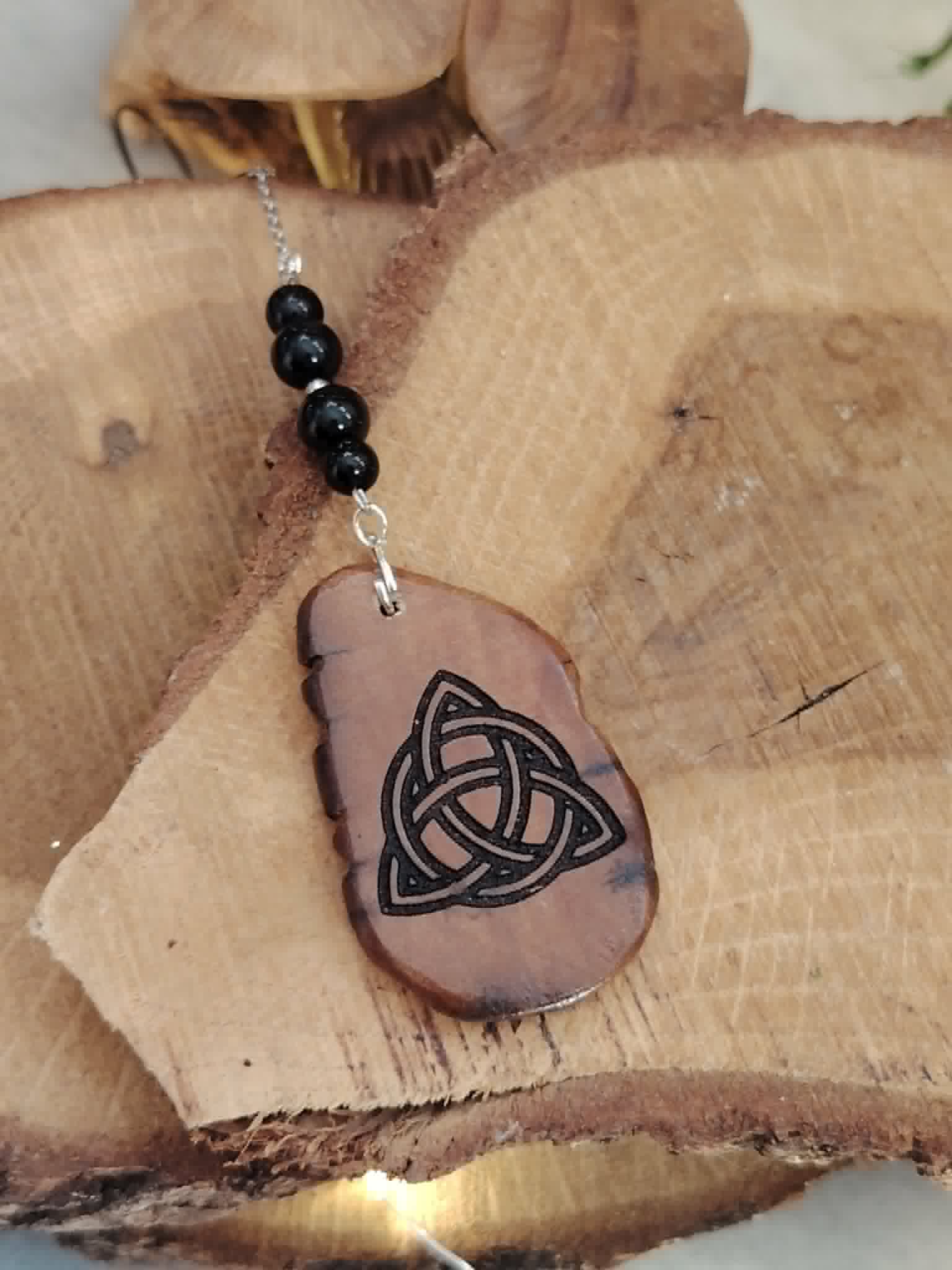 Face Triquetra du bijou de rétroviseur Magic Gaïa sur rondins de bois, gravure celtique et perles de tourmaline noire pour équilibre.