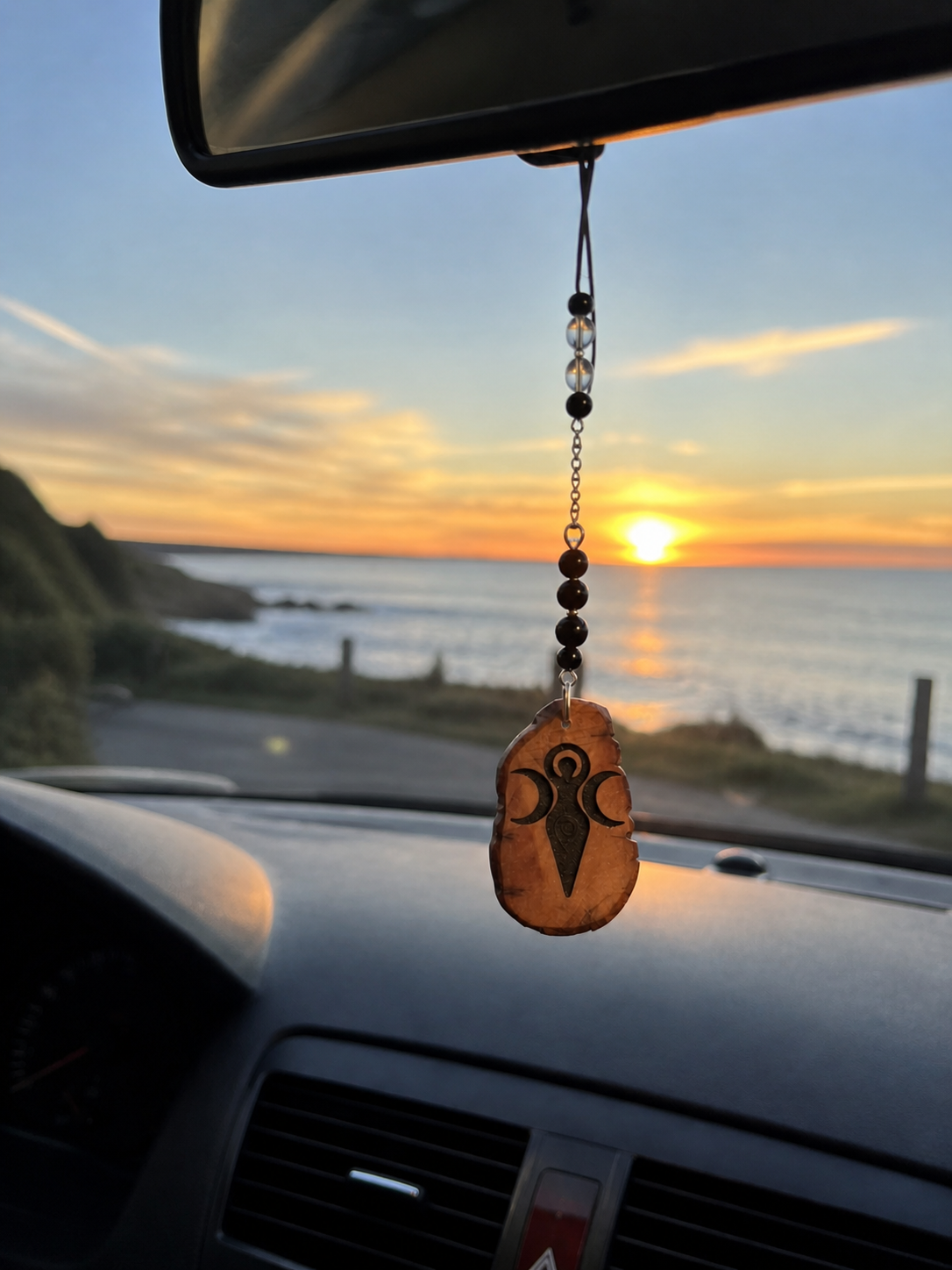 Bijou de rétroviseur Magic Gaïa suspendu dans une voiture face à la mer au coucher du soleil, symbole Gaïa pour voyager avec sérénité.