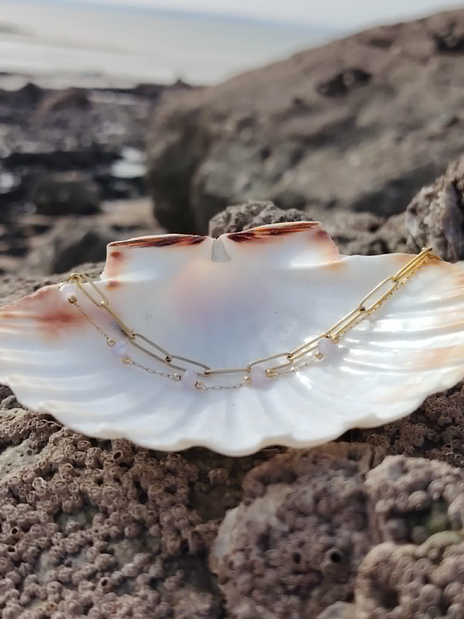 Bracelet doré avec perles violettes facettées posé dans un coquillage en bord de mer, bijou fin et lumineux.