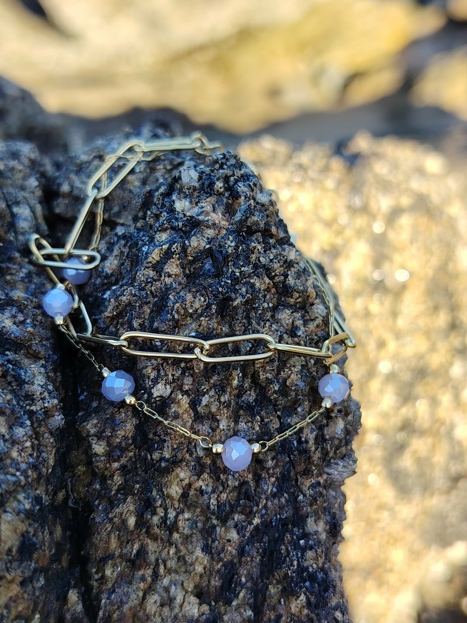 Bracelet doré à perles lavande sur roche côtière, bijou délicat pour une allure douce et féminine.