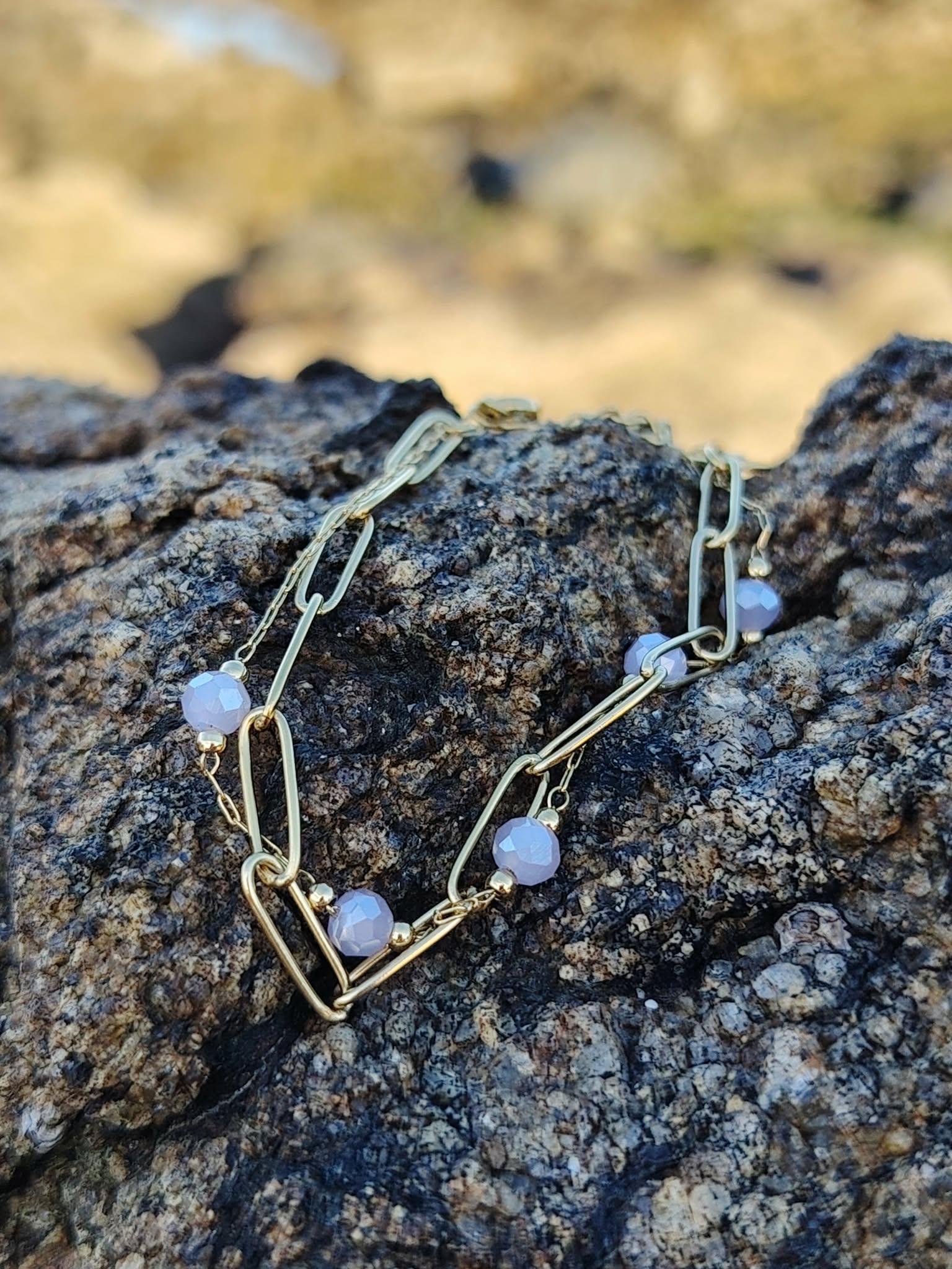 Gros plan du bracelet doré sur roche bretonne, maillons fins et perles violettes facettées pour une touche élégante.
