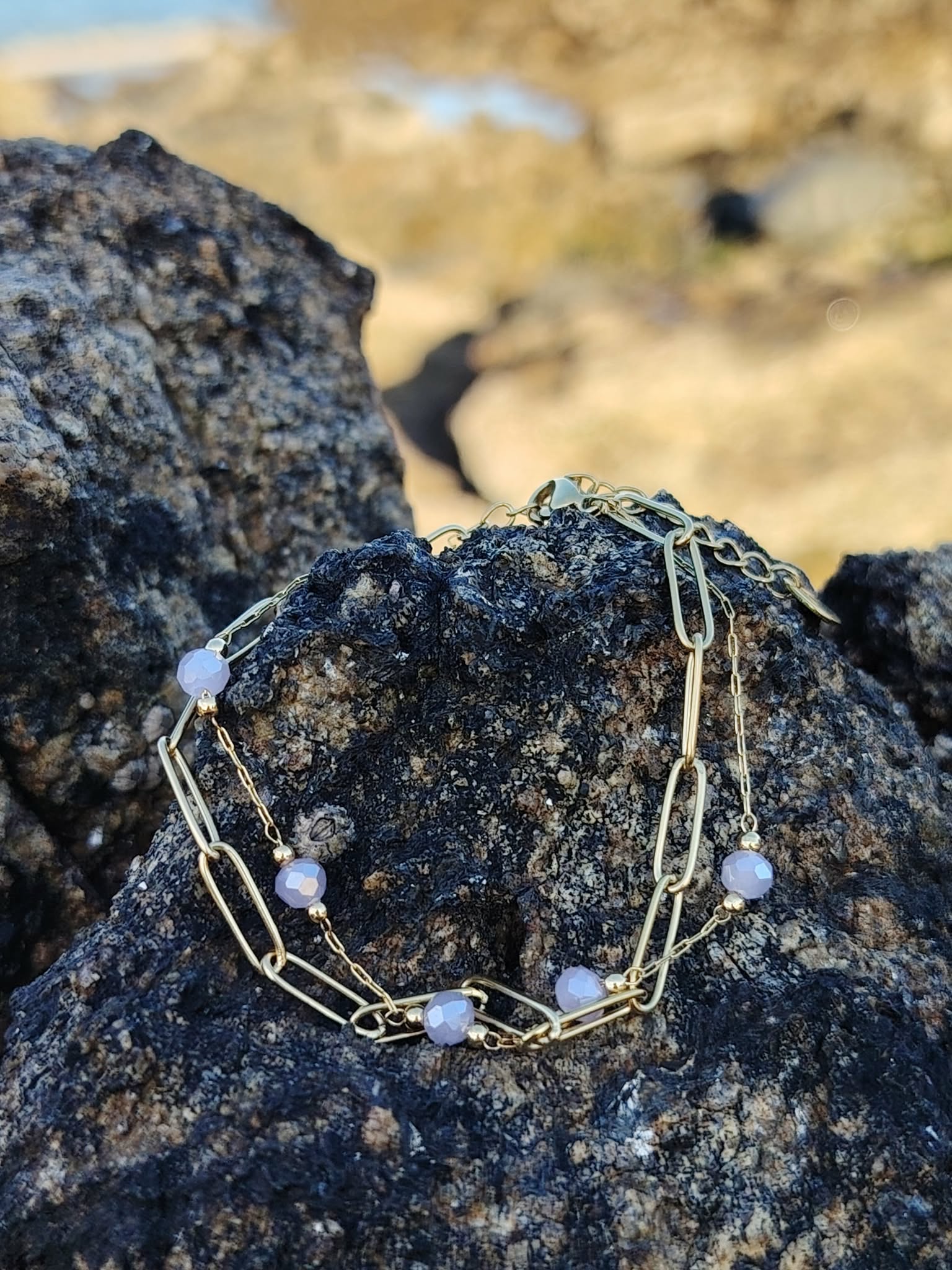 Bracelet doré posé sur pierre naturelle en bord de mer, perles violettes lumineuses pour élégance et sérénité.