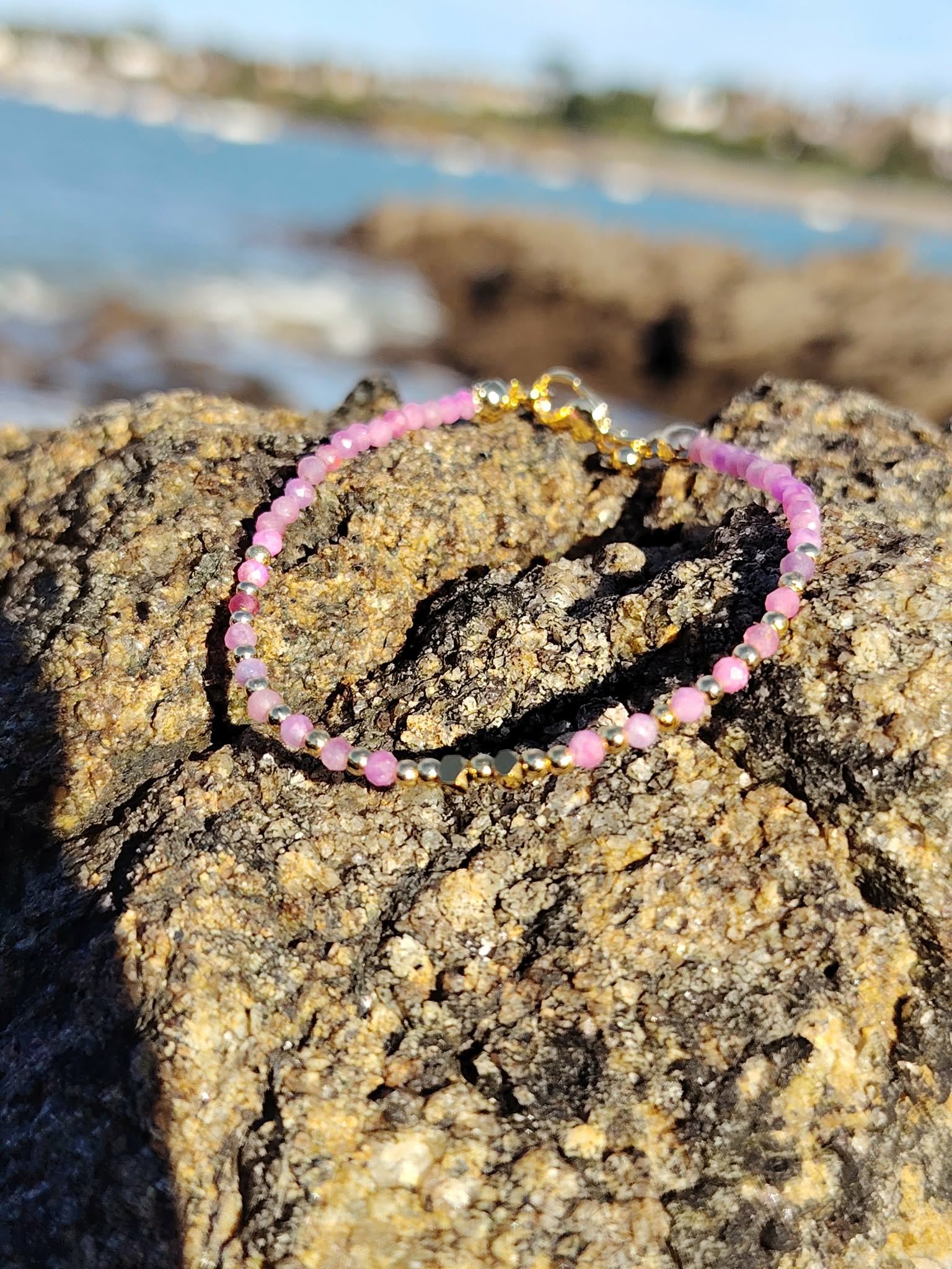 Bracelet Tourmaline rose facettée sur roche en Bretagne, cuivre plaqué or et mer floutée pour une allure lumineuse.