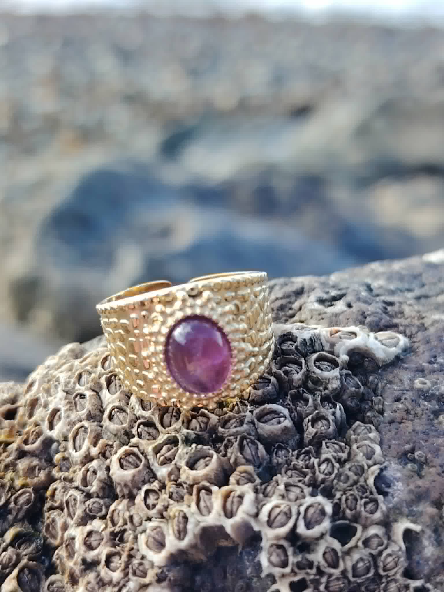 Bague dorée ajustable Améthyste sur roche de bord de mer en Bretagne, acier inoxydable texturé et pierre violette lumineuse.