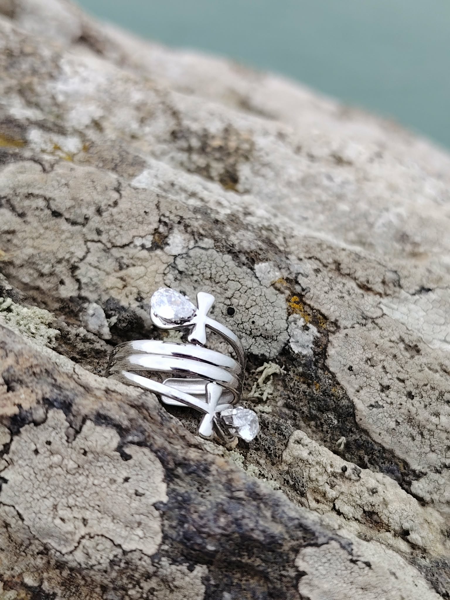 Bague croix de vie stylisée en acier inoxydable sur rocher, mer floutée en arrière-plan et strass goutte lumineux.