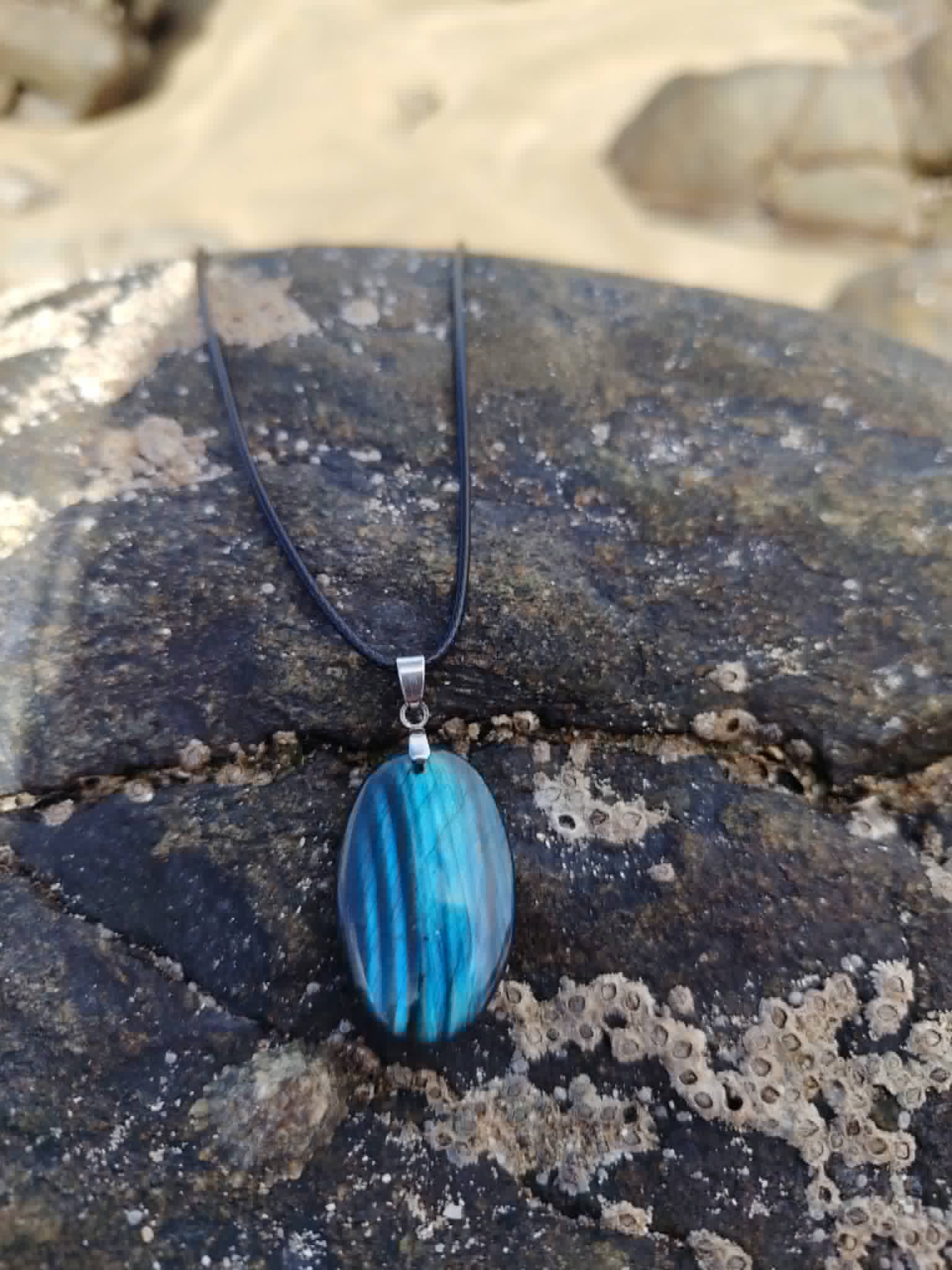 Pendentif Labradorite bleue sur roche bretonne avec sable en arrière-plan, cordon noir pour stabilité et protection.