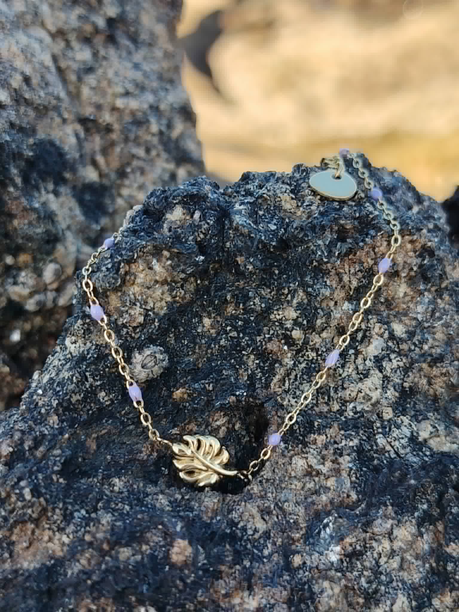 Bracelet Améthyste feuille dorée sur rocher sombre en bord de mer breton, chaîne fine en acier inoxydable et perles violettes pour sérénité, intuition et équilibre émotionnel.