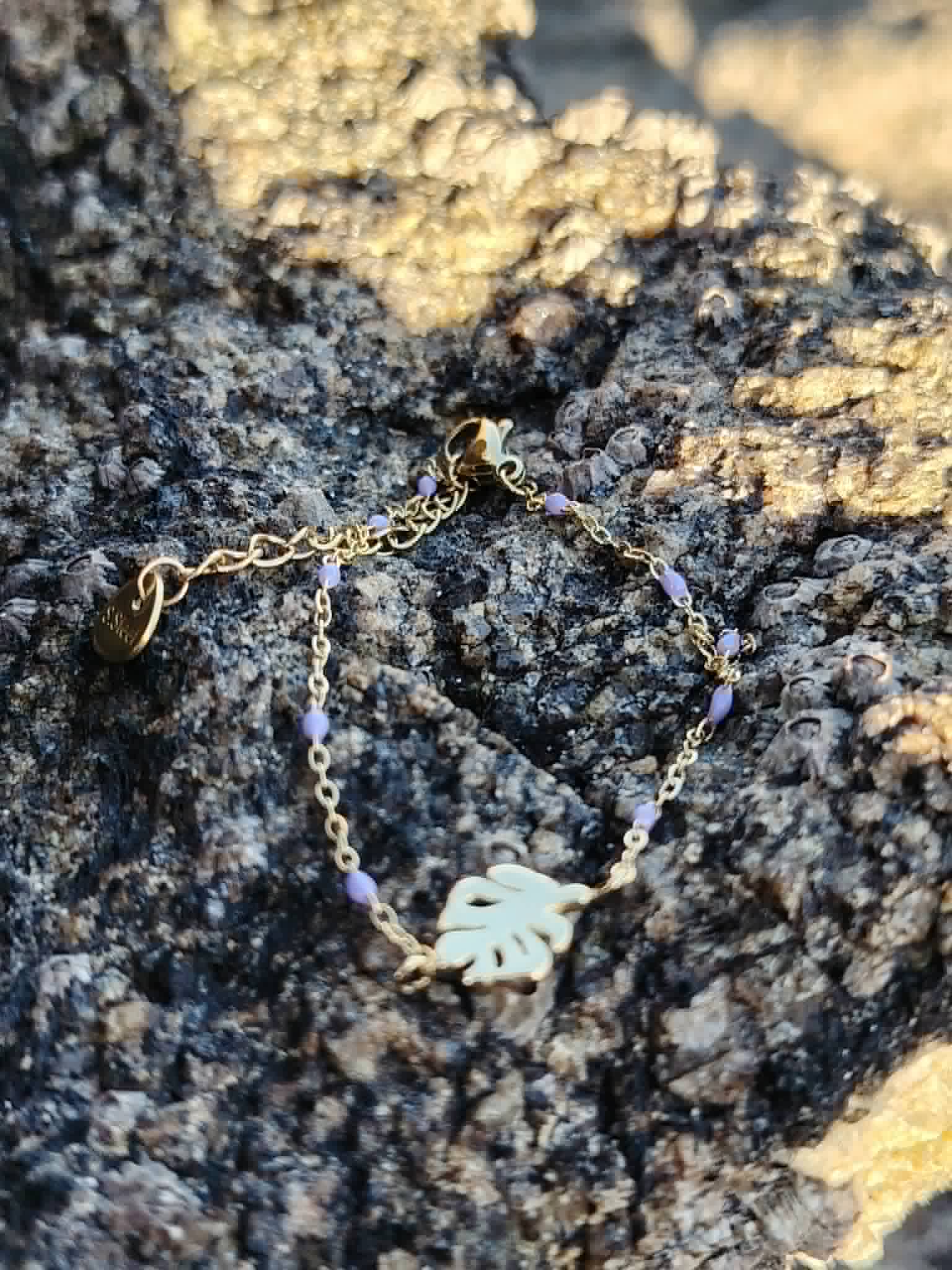 Bracelet Améthyste feuille dorée posé sur rocher coquillé en bord de mer, rallonge et médaillon visibles, perles violettes pour paix intérieure, méditation et sensibilité.