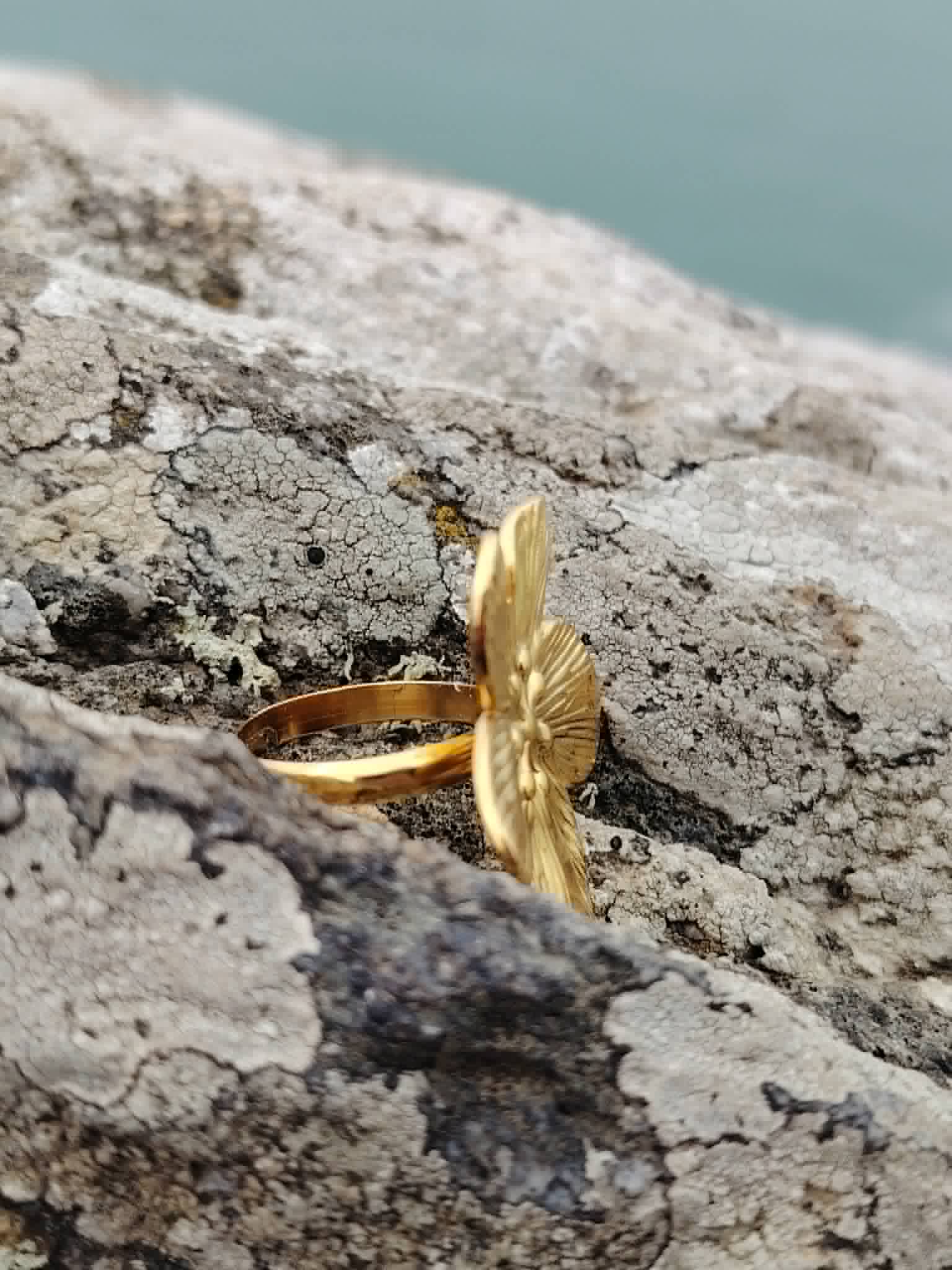 Bague fleur dorée réglable vue de profil sur rocher en bord de mer, anneau ajustable visible et fleur texturée en acier inoxydable.