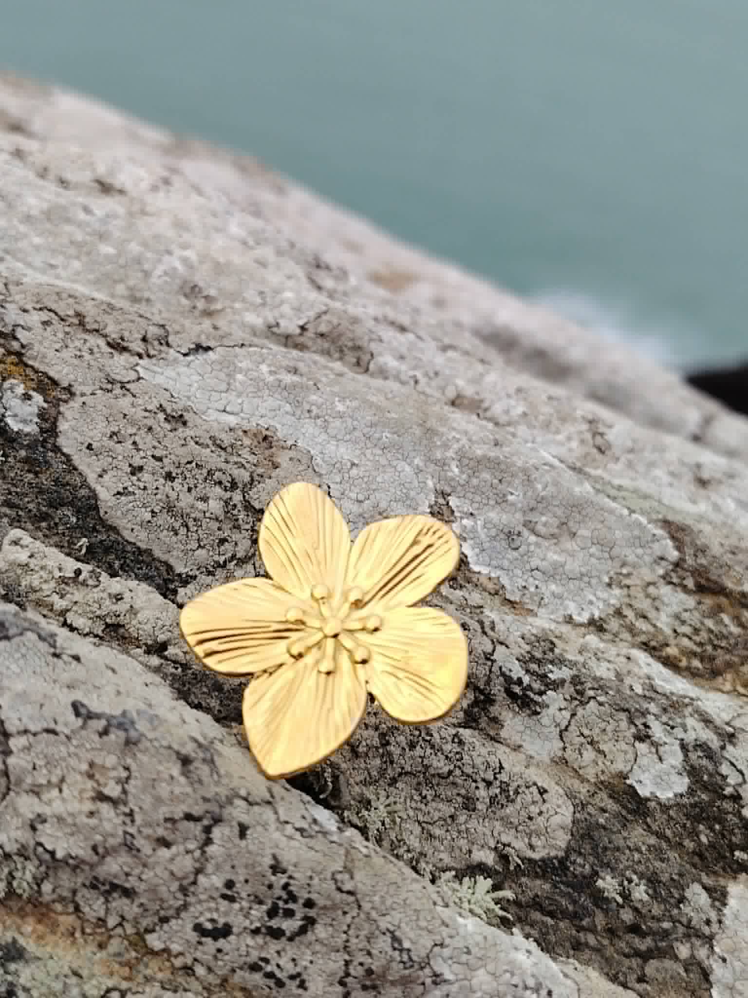 Bague fleur dorée réglable en acier inoxydable sur rocher côtier breton, motif floral texturé et finition or 18K pour un style solaire.