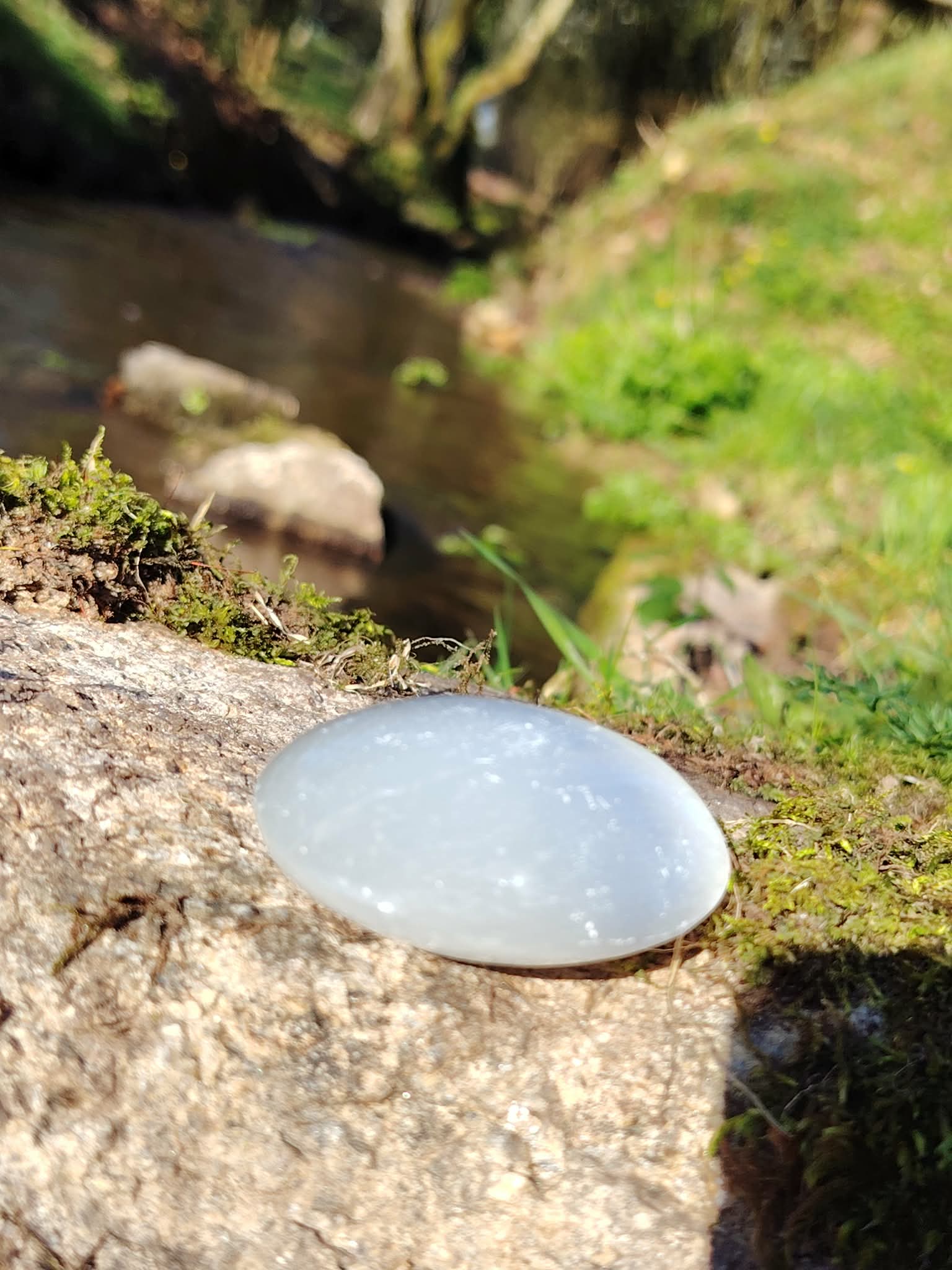 Galet Sélénite naturel posé au bord d’un ruisseau, pierre ovale lumineuse pour décoration spirituelle et moment de calme.