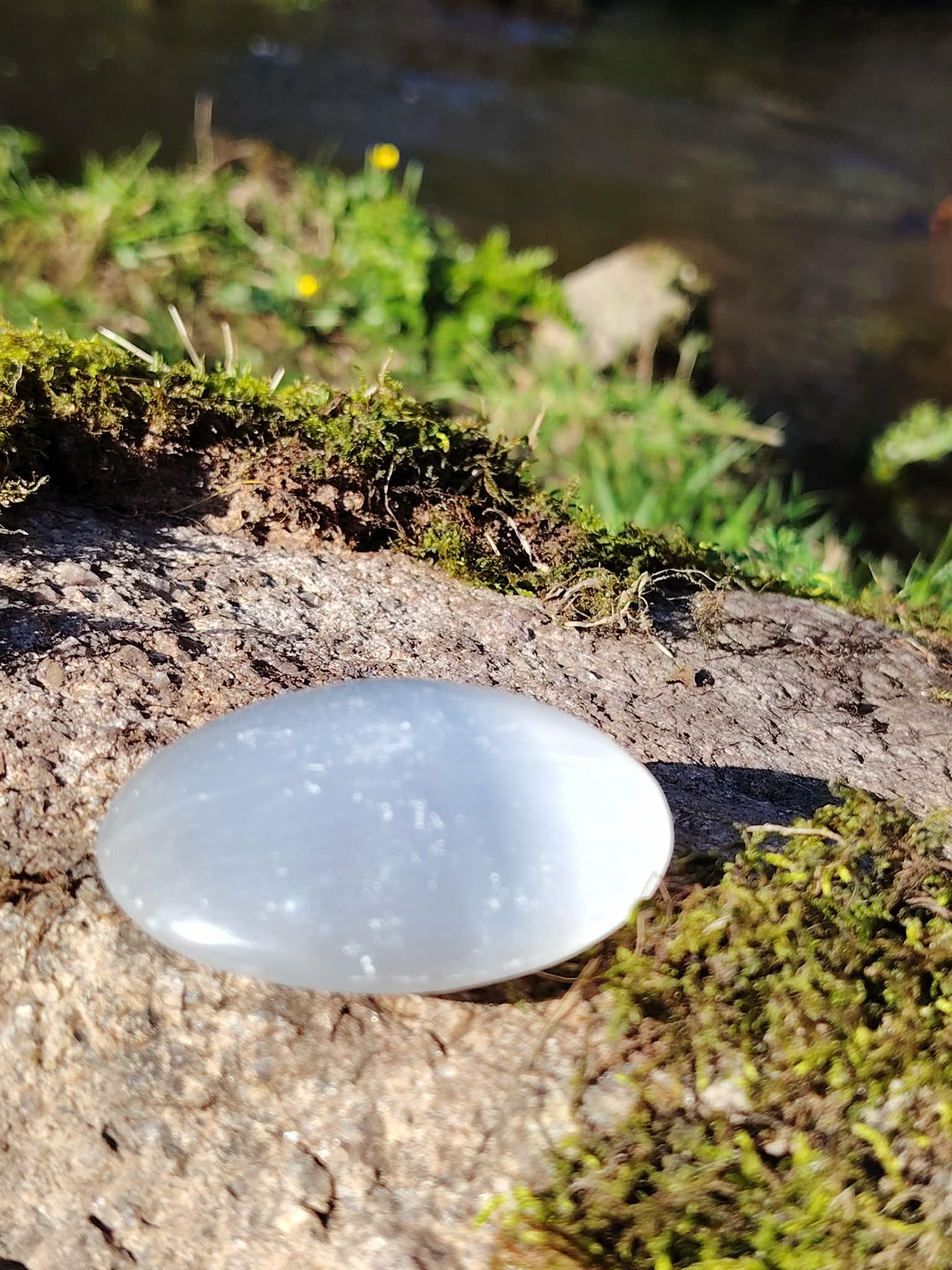 Galet Sélénite blanc poli posé sur rocher moussu près d’un ruisseau, pierre lumineuse pour purification douce et clarté intérieure.