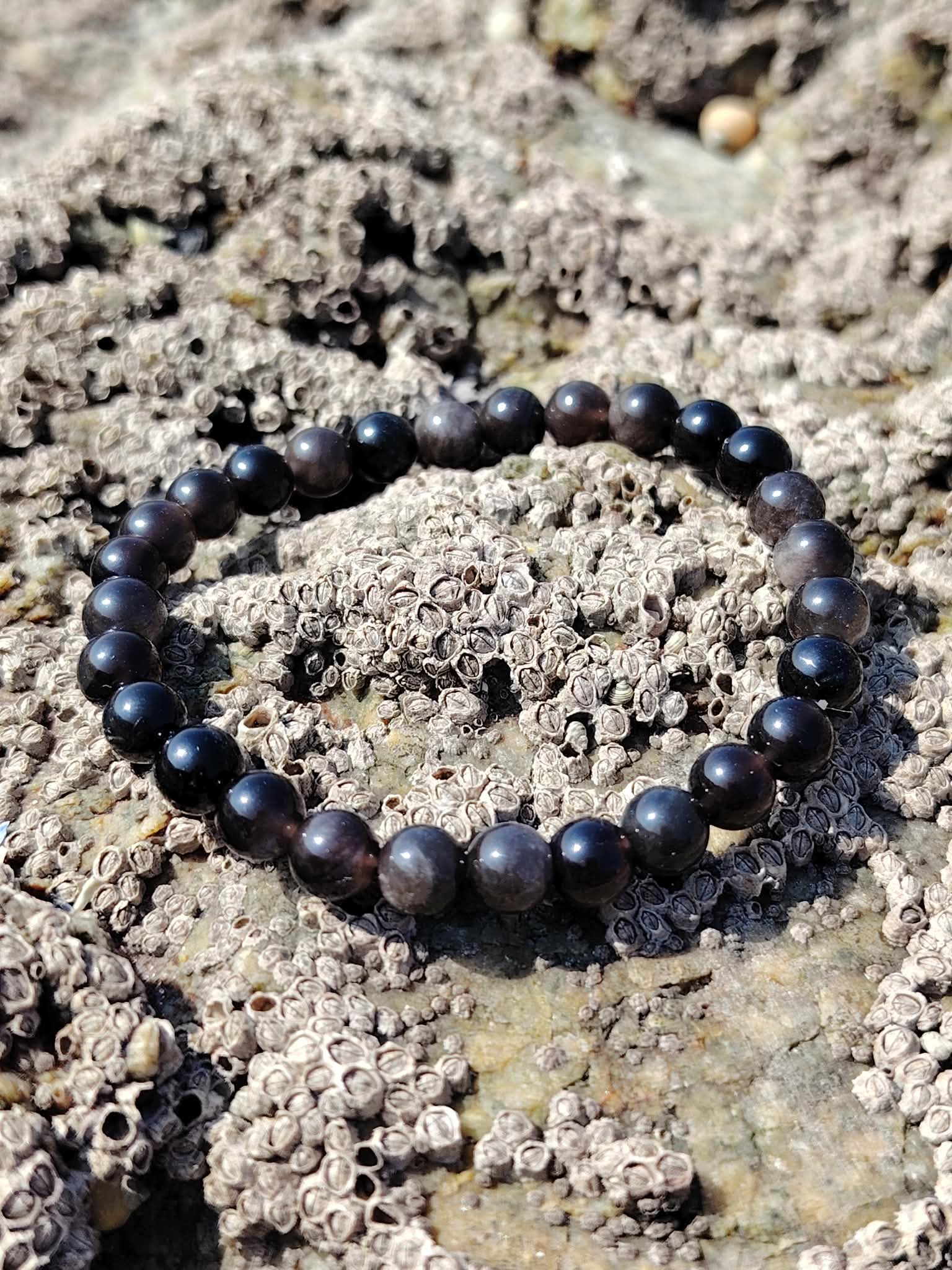 Bracelet Obsidienne Argentée posé sur roche texturée en Bretagne, perles foncées lumineuses pour soutenir les limites personnelles et la force intérieure.