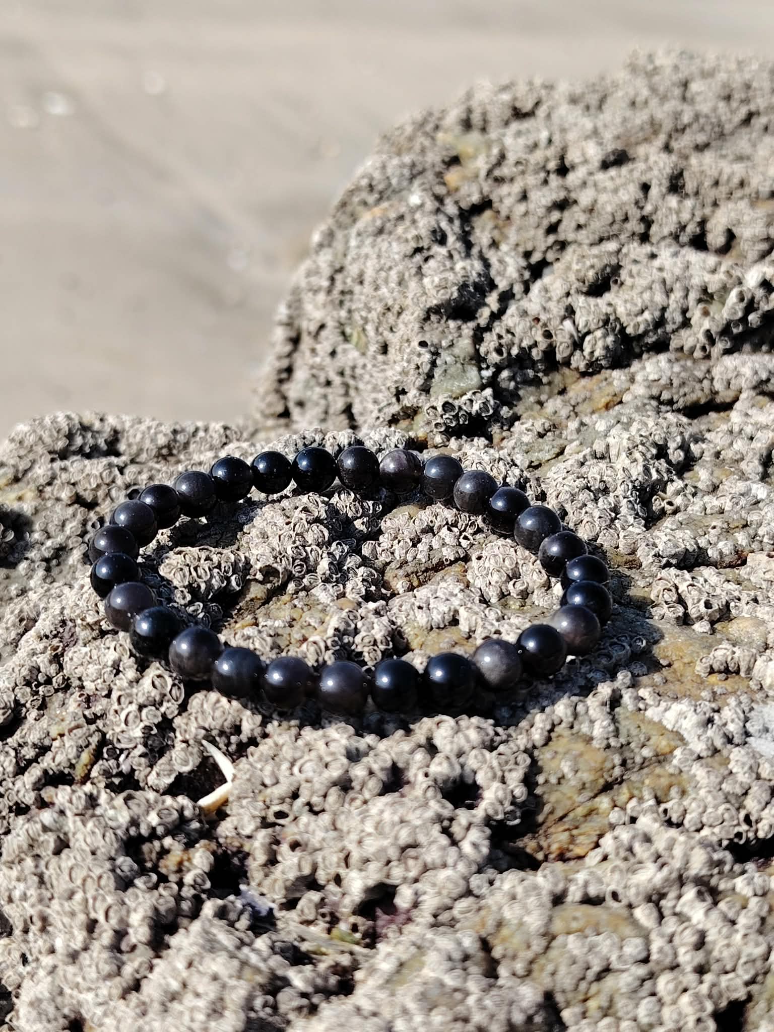 Bracelet Obsidienne Argentée en perles rondes sur rochers côtiers, bijou sobre aux reflets fumés pour transformation intérieure et recentrage.