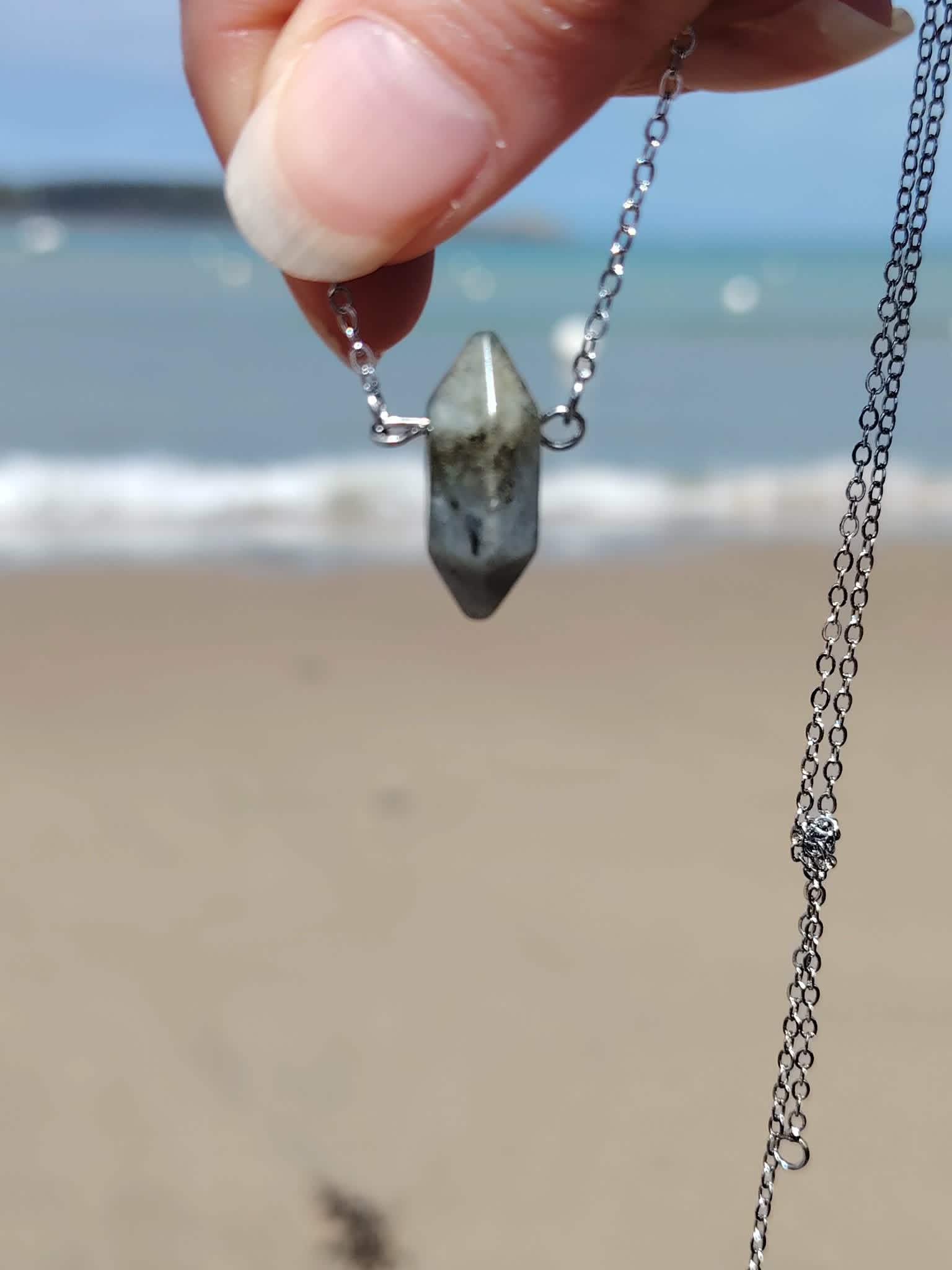 Pendentif pointe Labradorite tenu devant la mer, chaîne argentée en acier inoxydable et pierre naturelle aux reflets discrets.