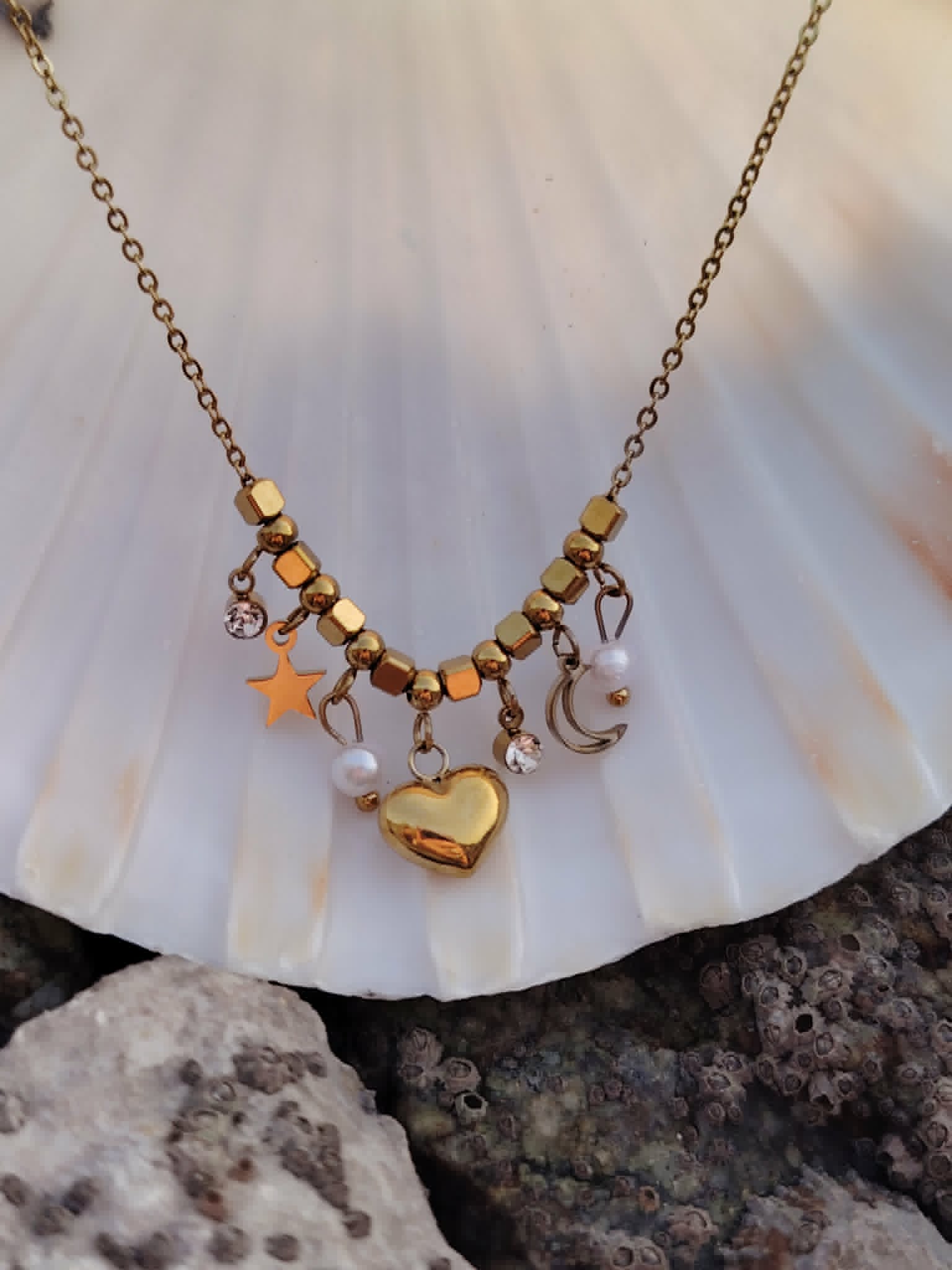 Collier doré cœur lune étoile posé sur coquillage blanc et rochers, bijou acier inoxydable avec perles blanches et strass lumineux.