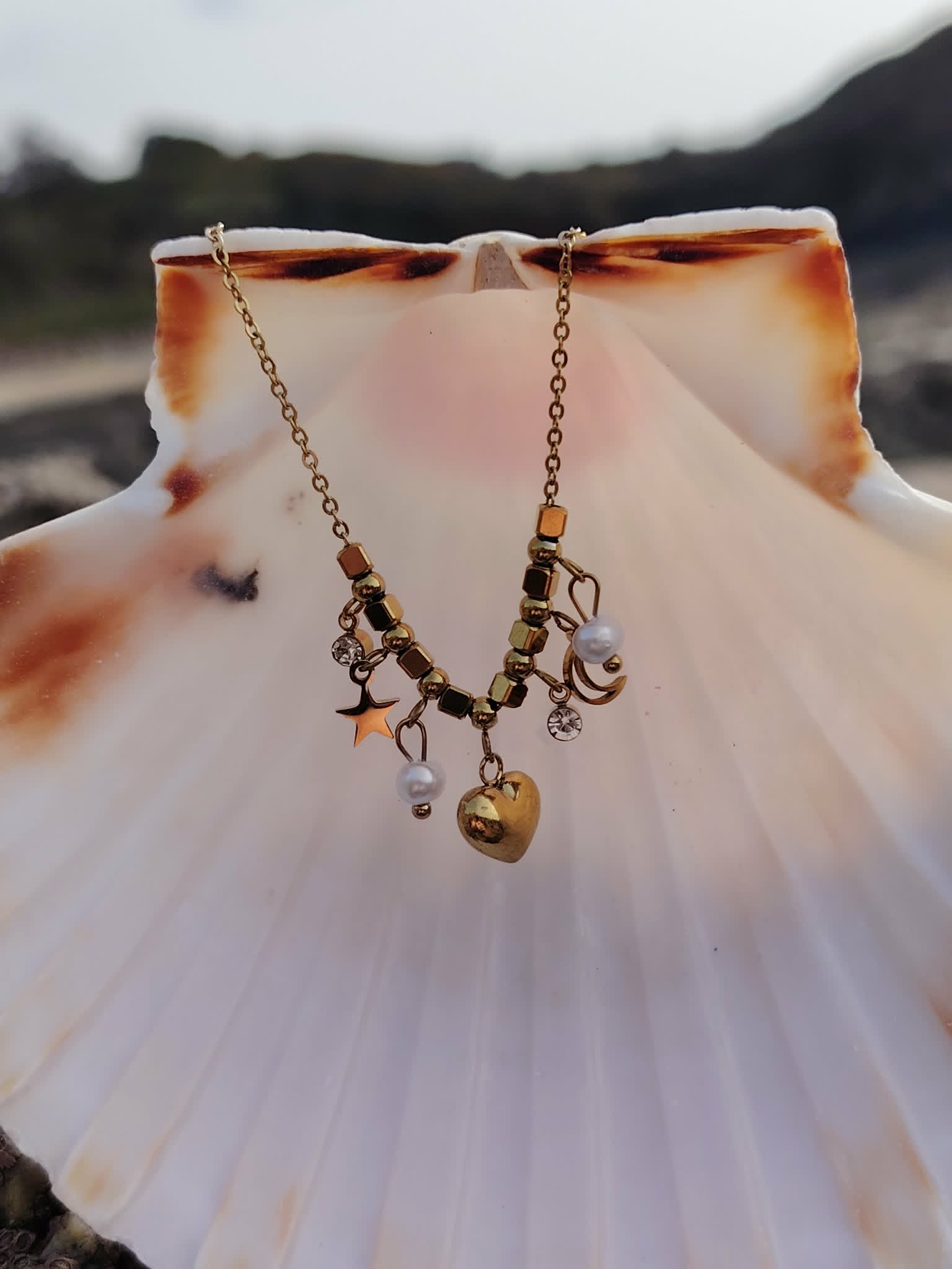 Collier doré à breloques célestes suspendu sur coquillage blanc, cœur central, lune, étoile et perles nacrées en bord de mer.