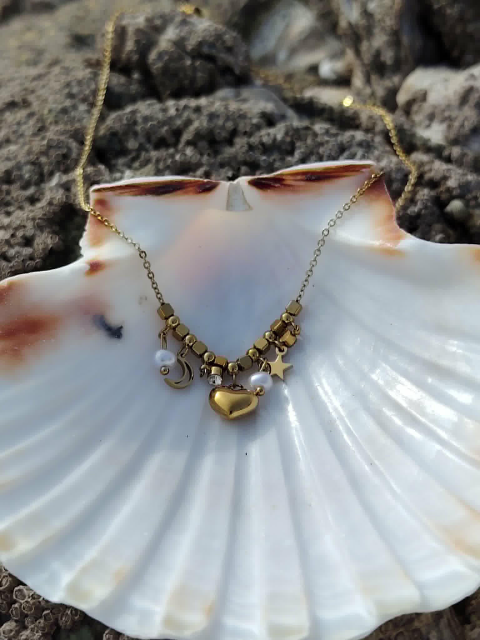 Collier acier inoxydable doré présenté sur grand coquillage blanc, breloques lune, étoile, cœur et perles dans un décor marin naturel.