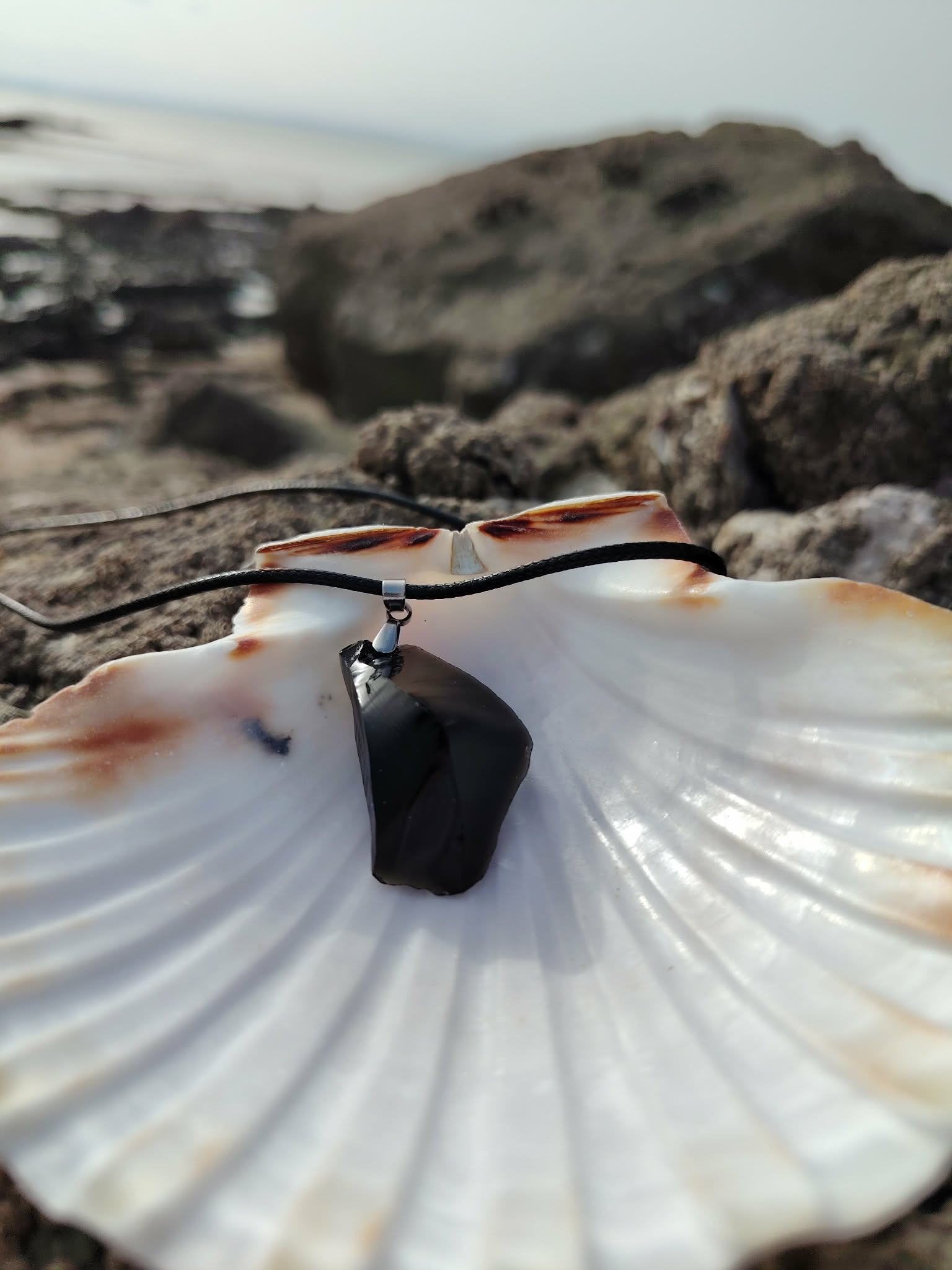 Collier Obsidienne noire sur coquillage face aux rochers bretons, pendentif sombre pour recentrage et énergie protectrice.