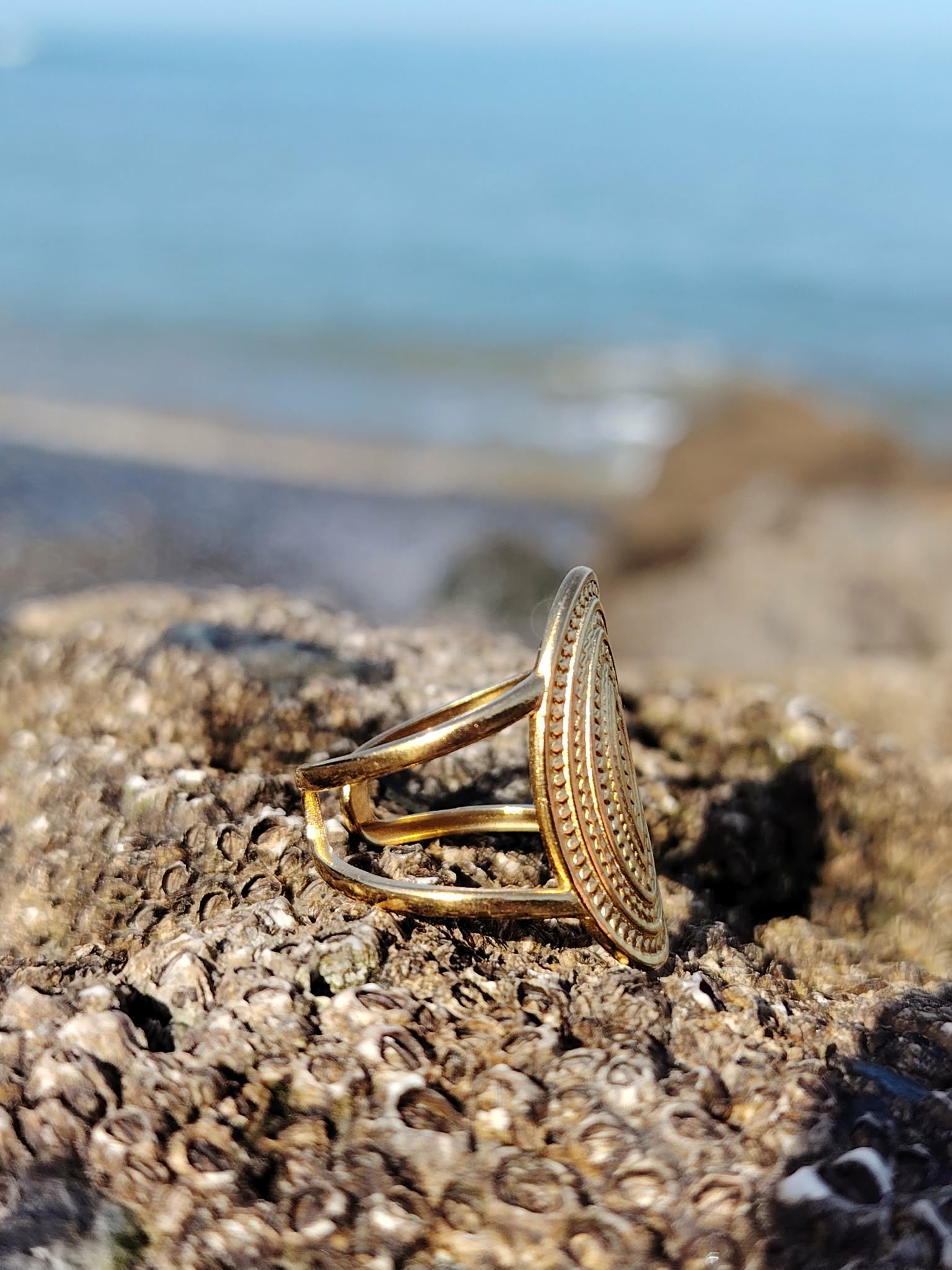 Bague ovale dorée vue de profil sur roche coquillée, anneau acier inoxydable et motif travaillé face au bord de mer.