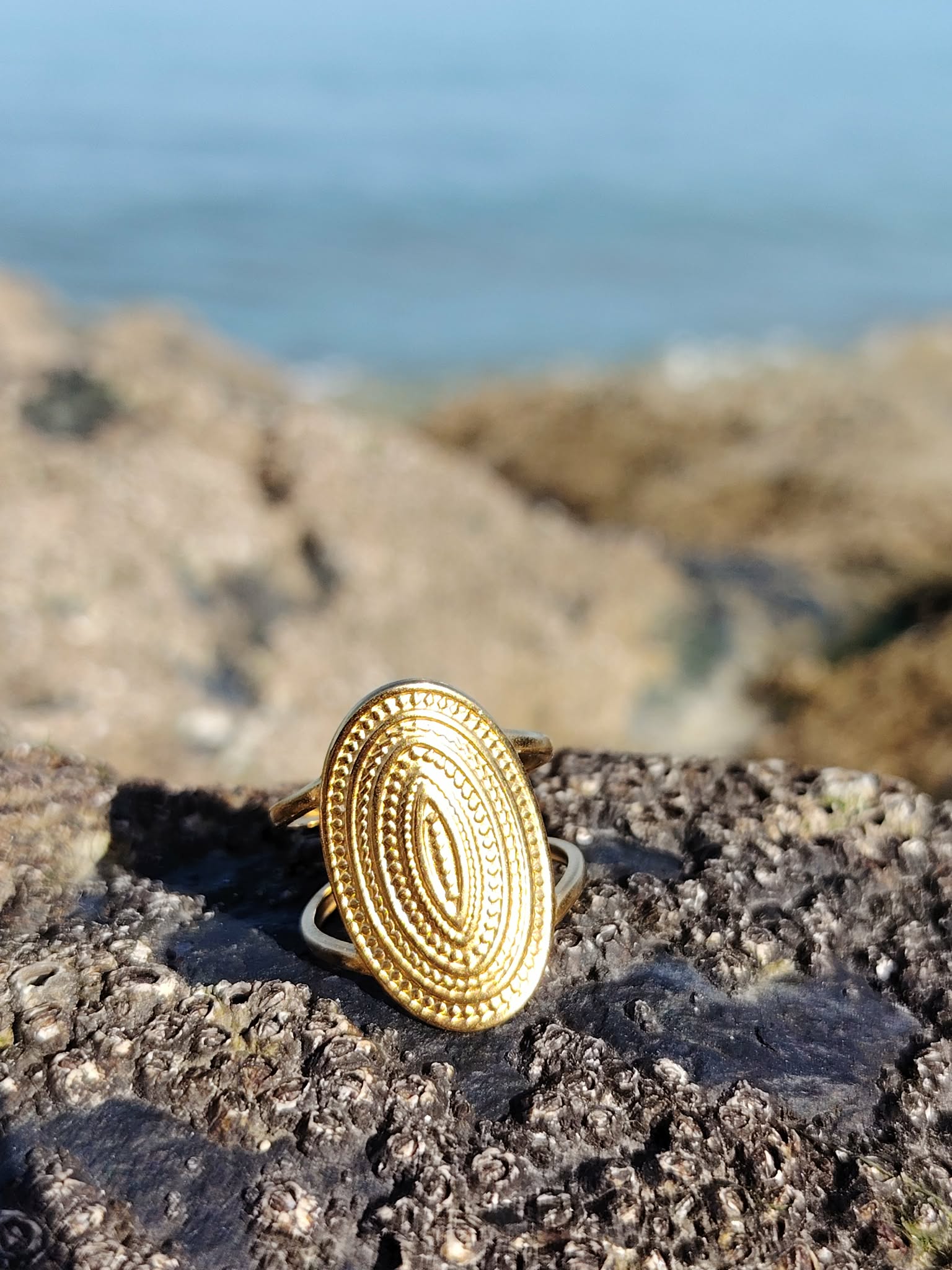 Bague dorée ovale texturée sur rocher en bord de mer, bijou acier inoxydable au style vintage, solaire et raffiné.