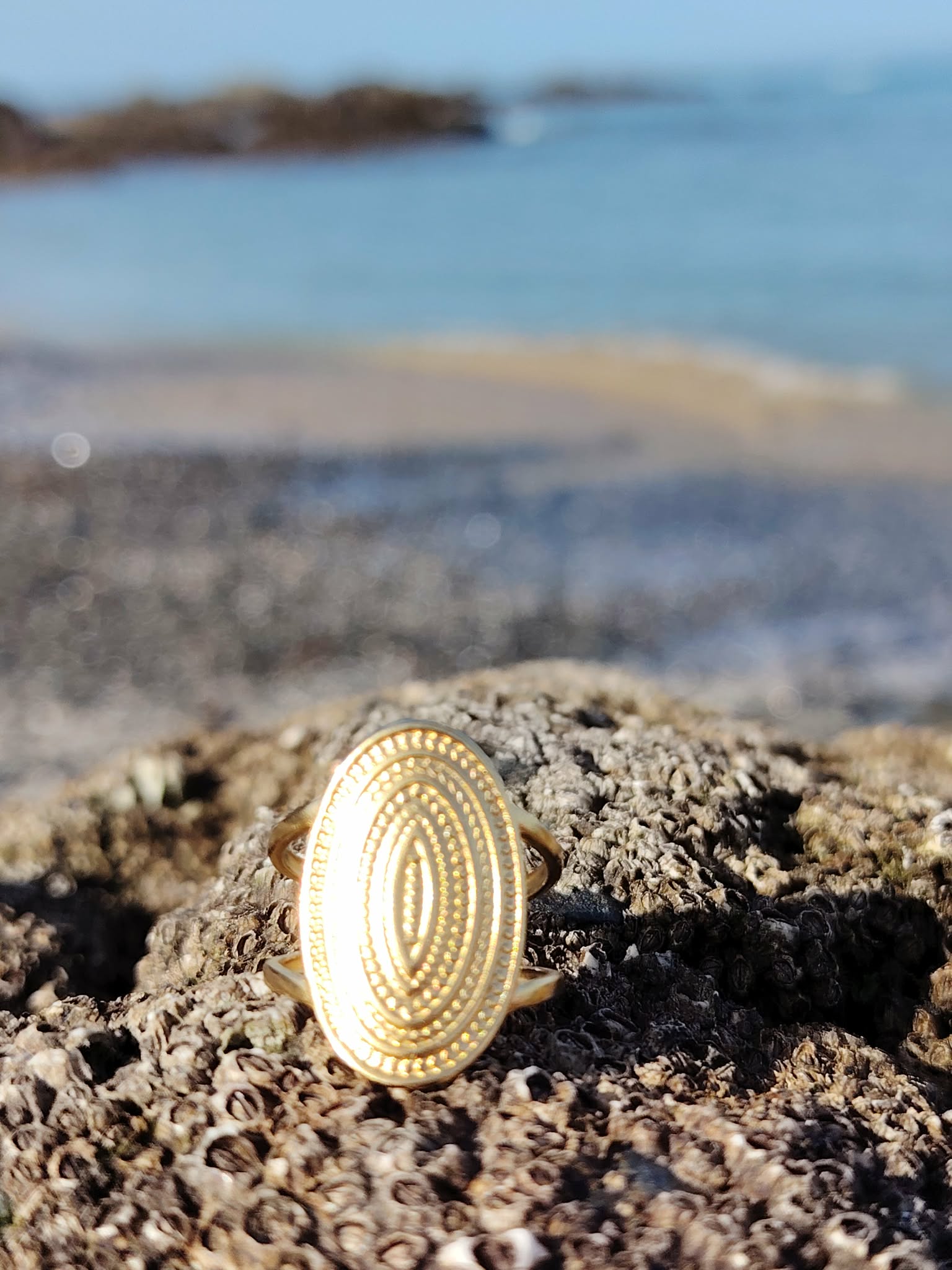 Bague dorée en acier inoxydable sur décor marin, plateau ovale texturé aux reflets lumineux et détails concentriques.