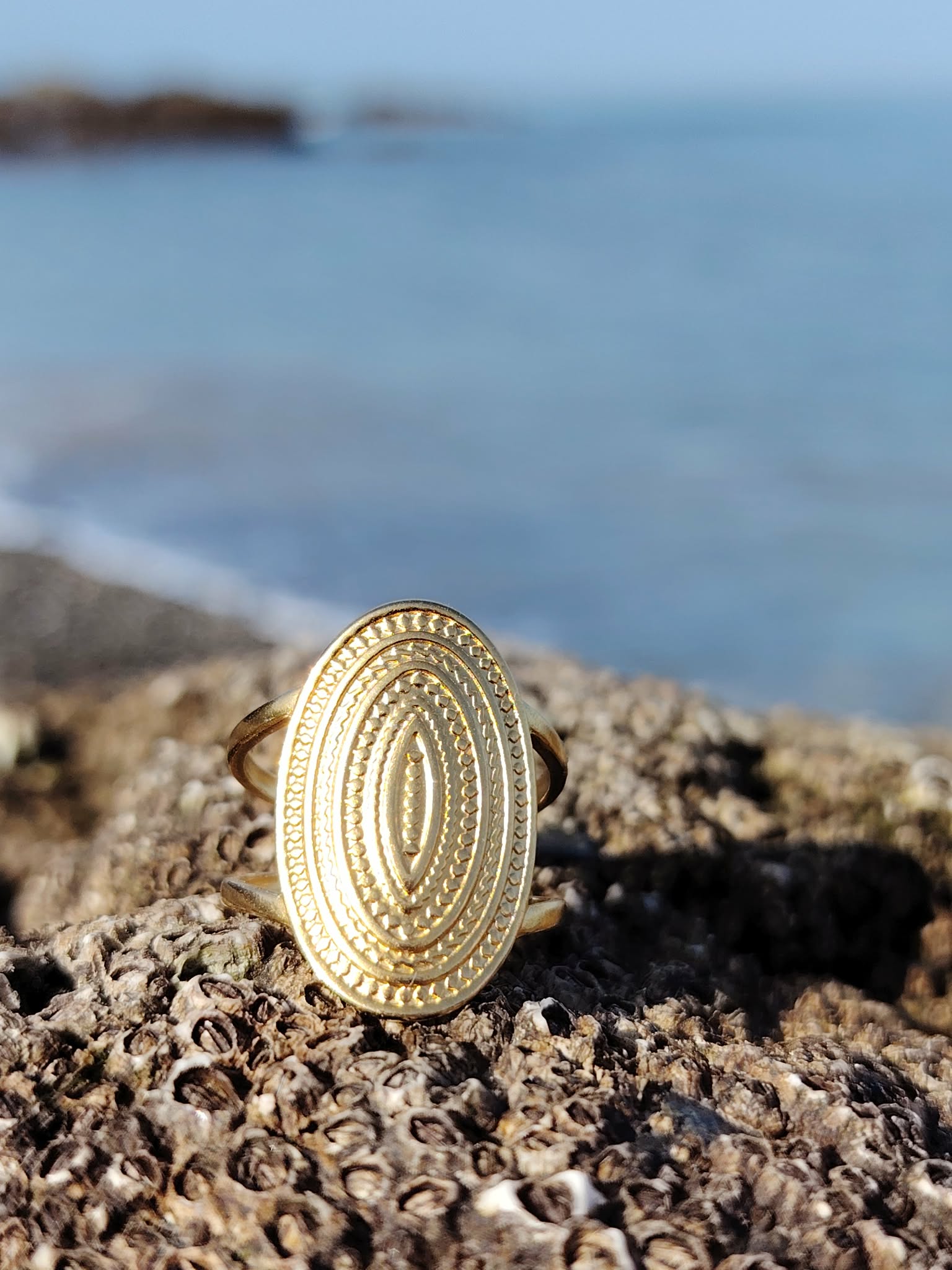 Bague dorée ovale texturée en acier inoxydable sur roche coquillée en bord de mer, motif relief lumineux et style bohème chic.