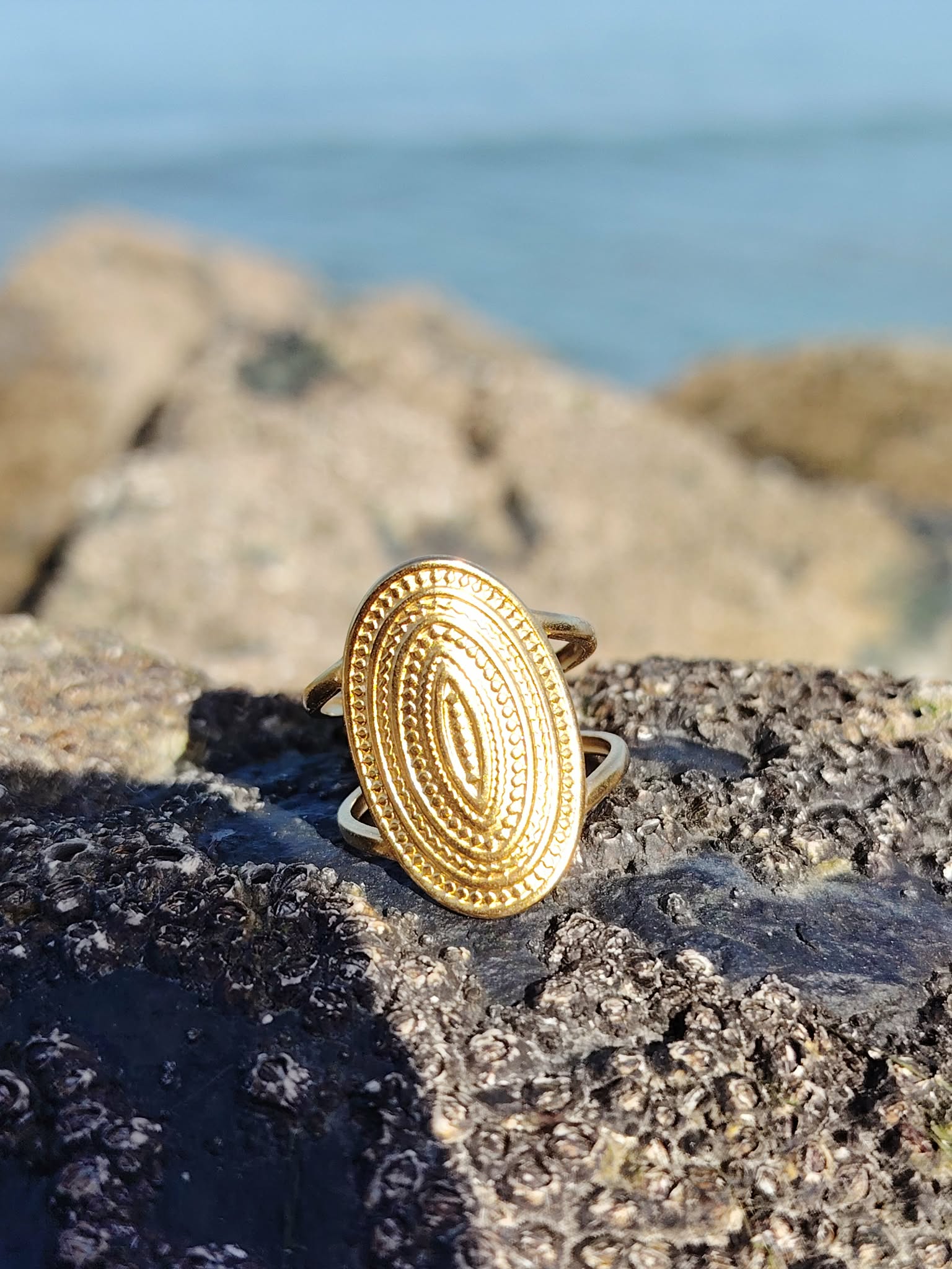 Bague dorée texturée posée sur rocher côtier, plateau ovale en relief et finition brillante pour une touche bohème chic.