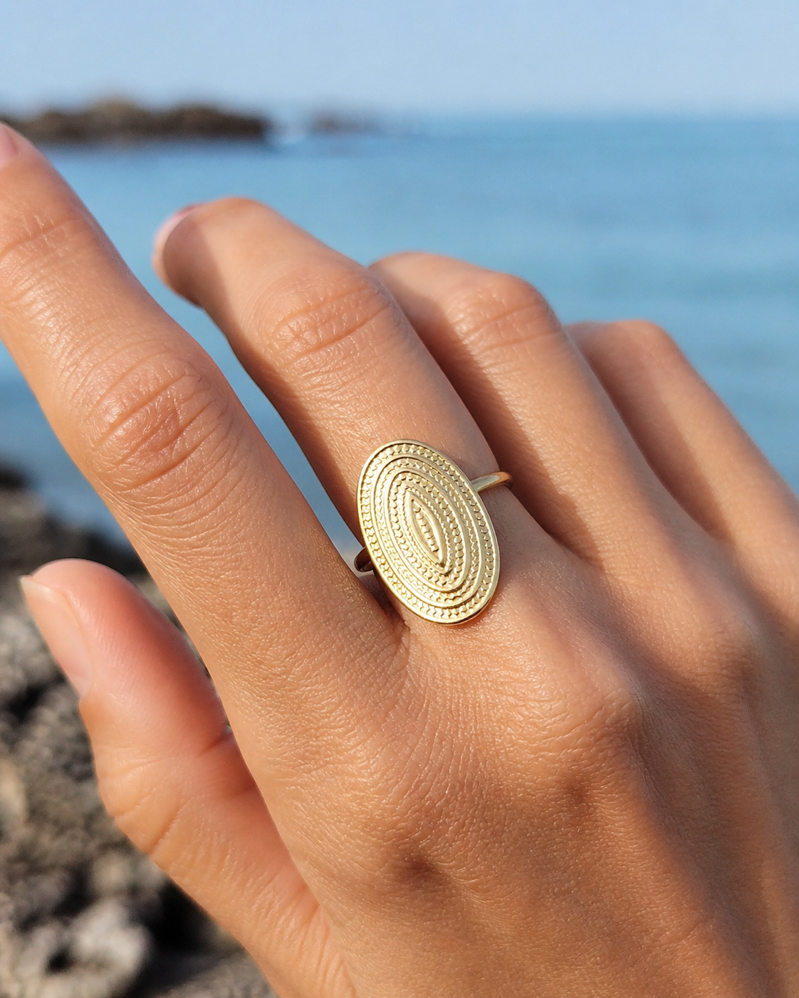 Bague dorée ovale portée au doigt face à la mer, acier inoxydable texturé au motif relief élégant et solaire.